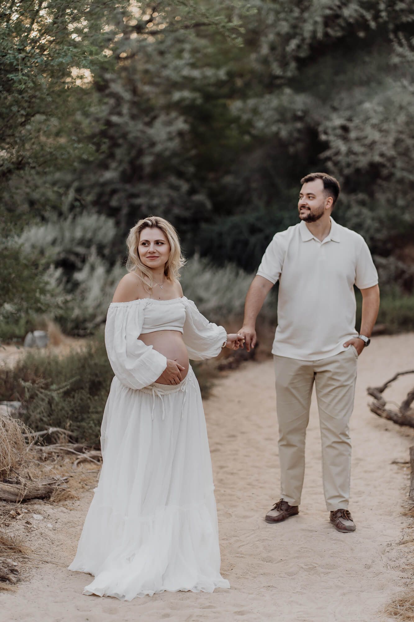 Romantic maternity photoshoot with couple in white and beige outfits on outdoor path.