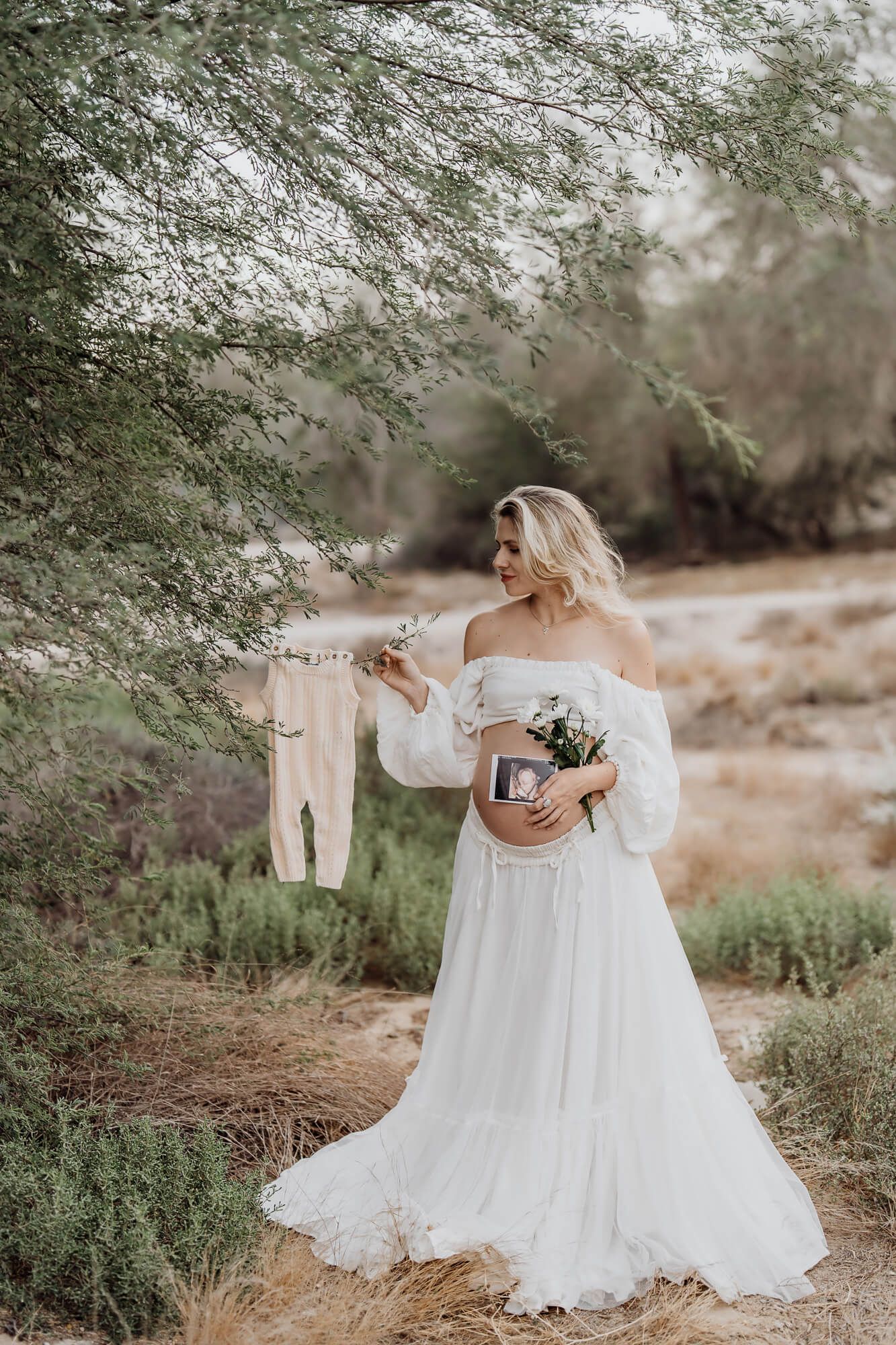 Woman in ethereal white dress holds tiny onesie and flower bouquet under desert trees.