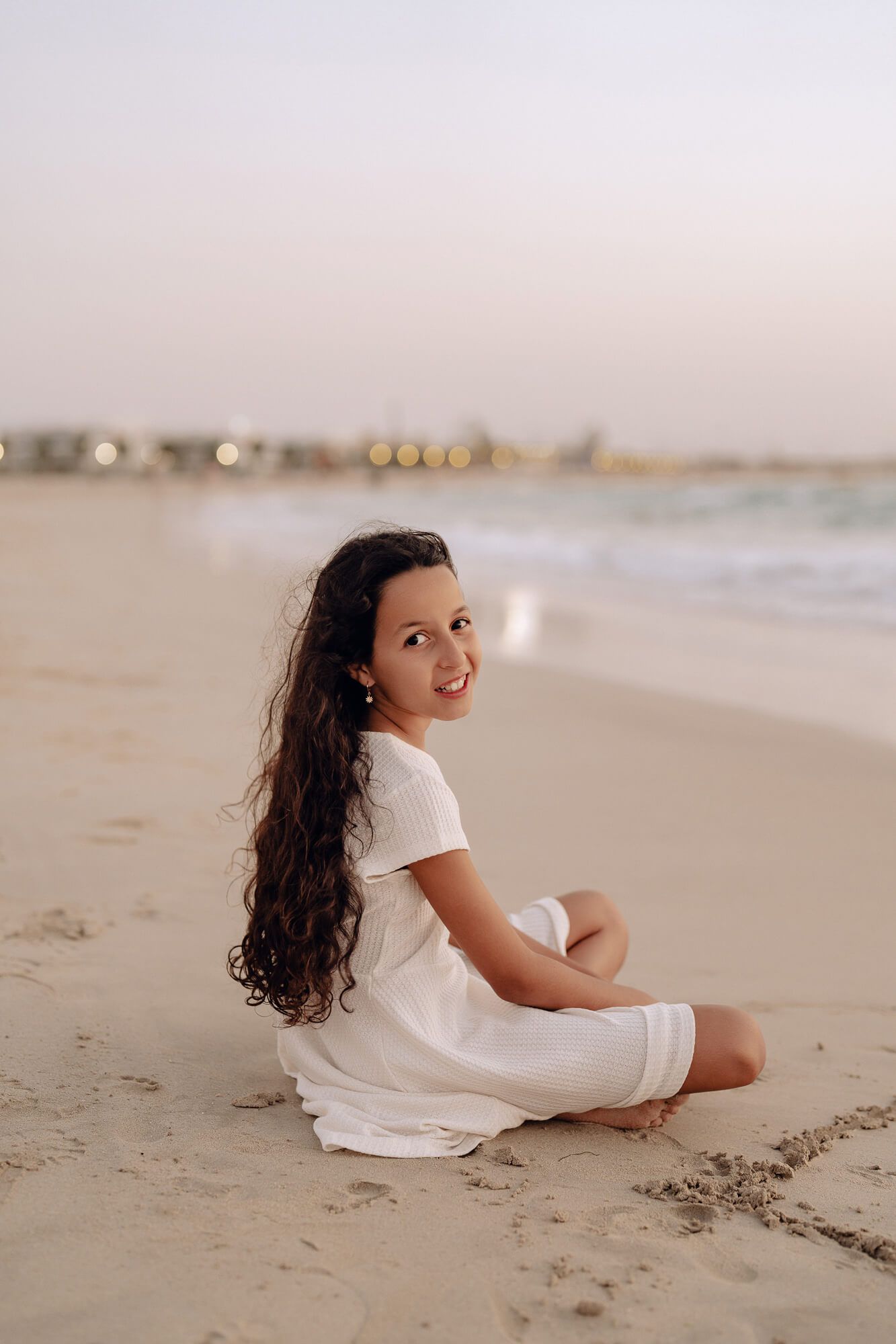 Person in white dress sitting on sandy beach during golden hour with waves in background.