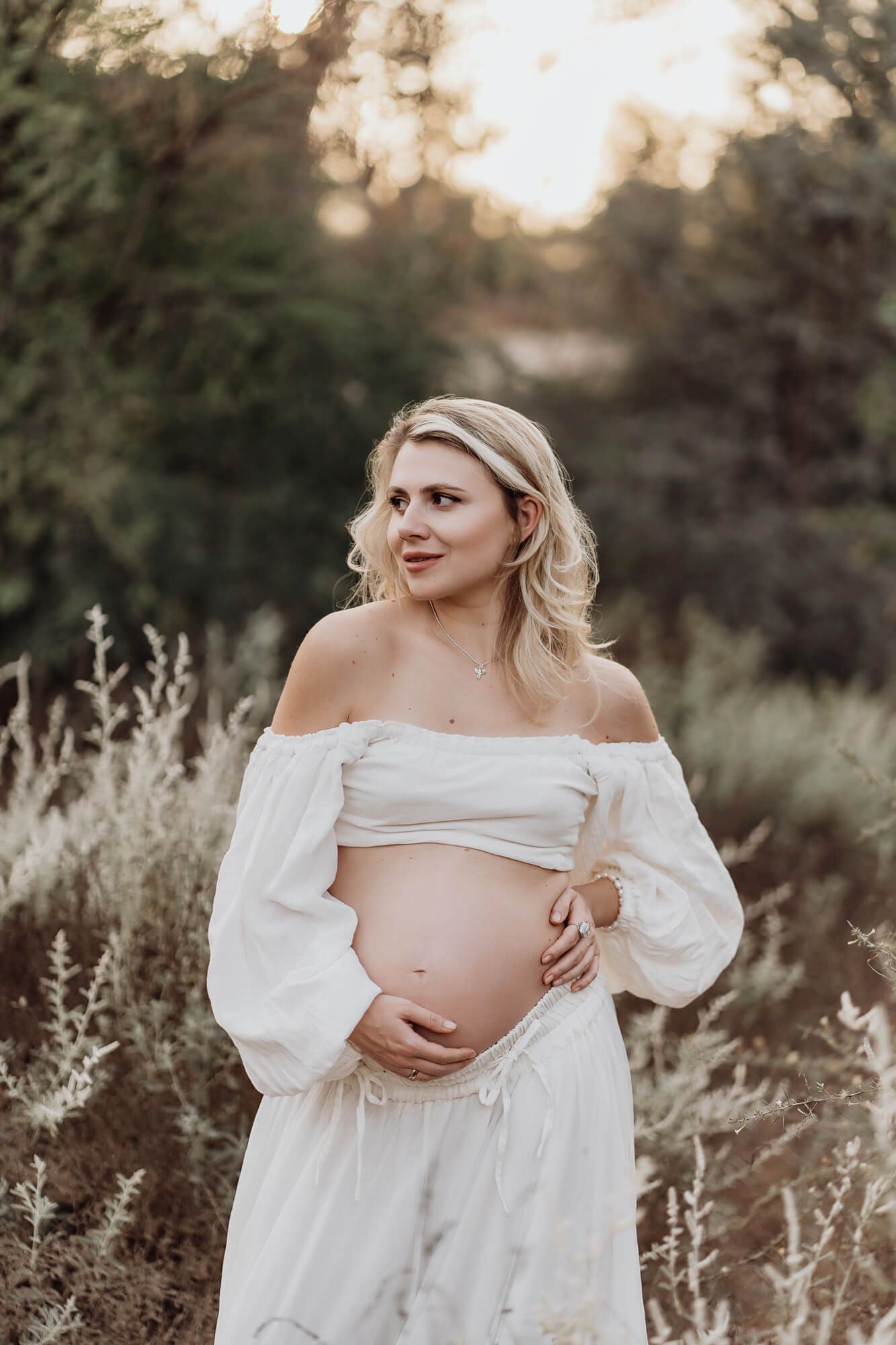 A maternity portrait of a woman in white off-shoulder dress against a sunset backdrop.