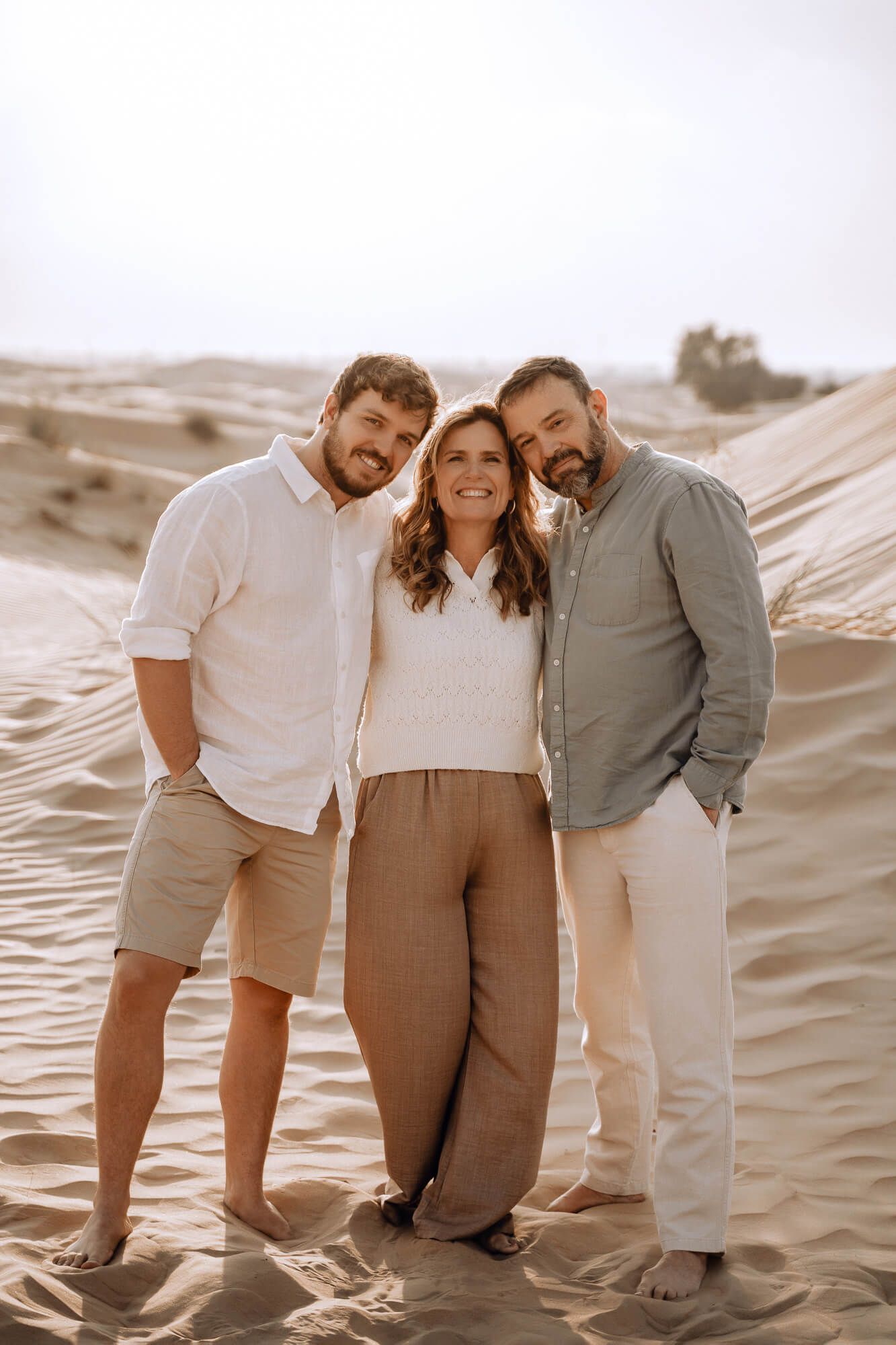 Three people sit together smiling on sand dunes in warm neutral toned outfits on a sunny day.