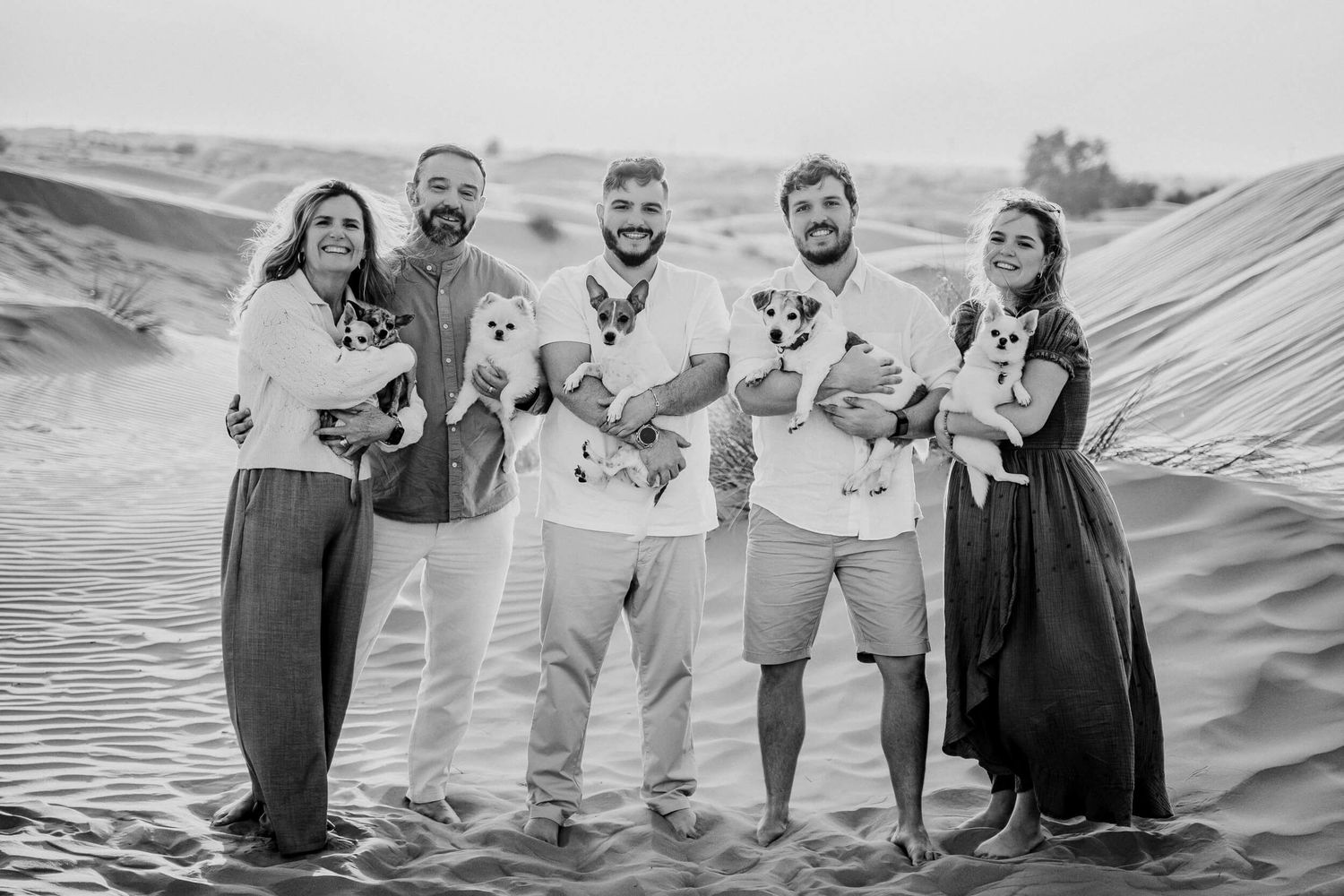 A group photo on sand dunes holding small dogs while wearing coordinated neutral-toned outfits.