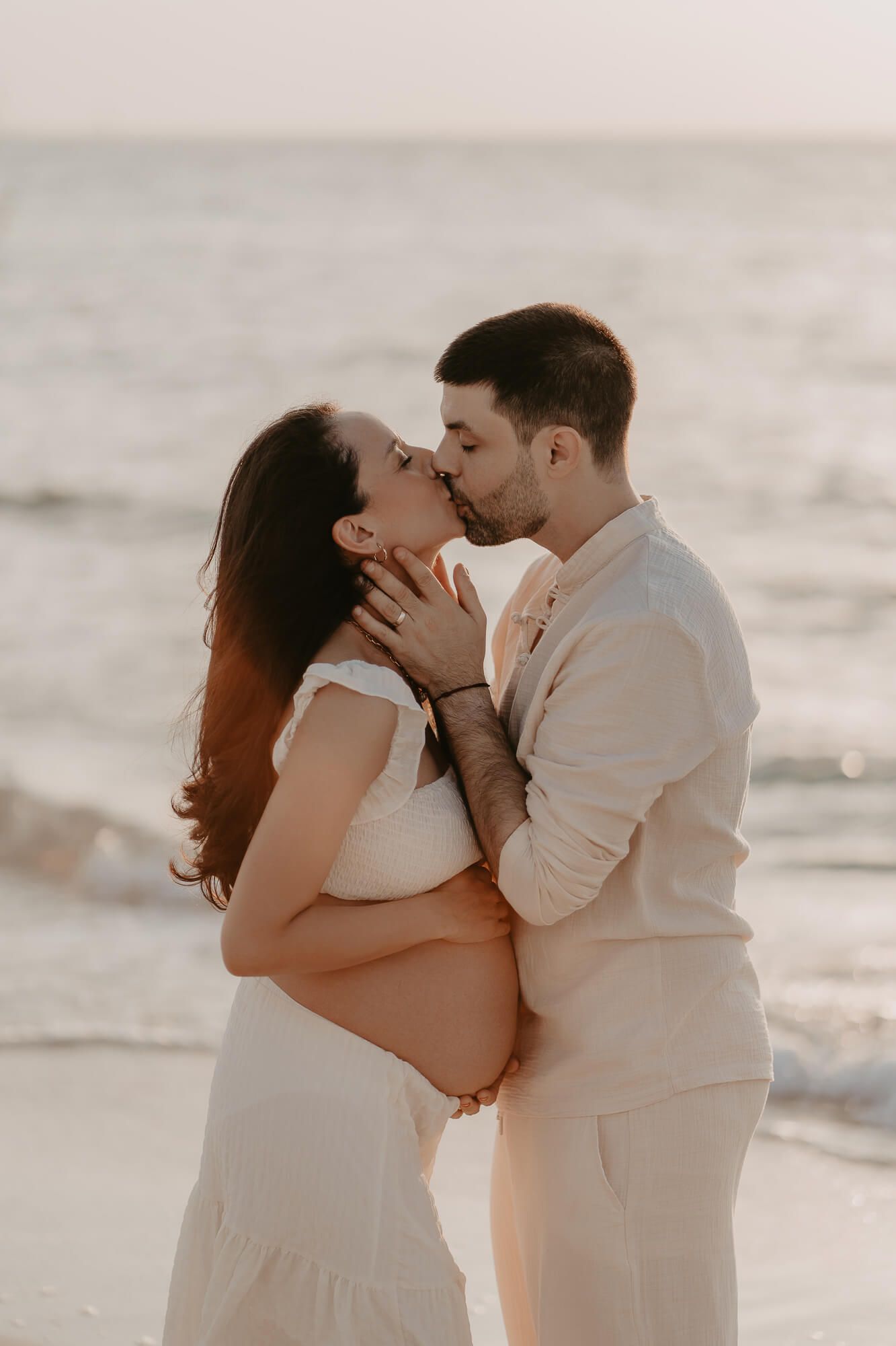 A romantic maternity photoshoot on the beach at sunset shows a tender moment between a couple in white outfits.