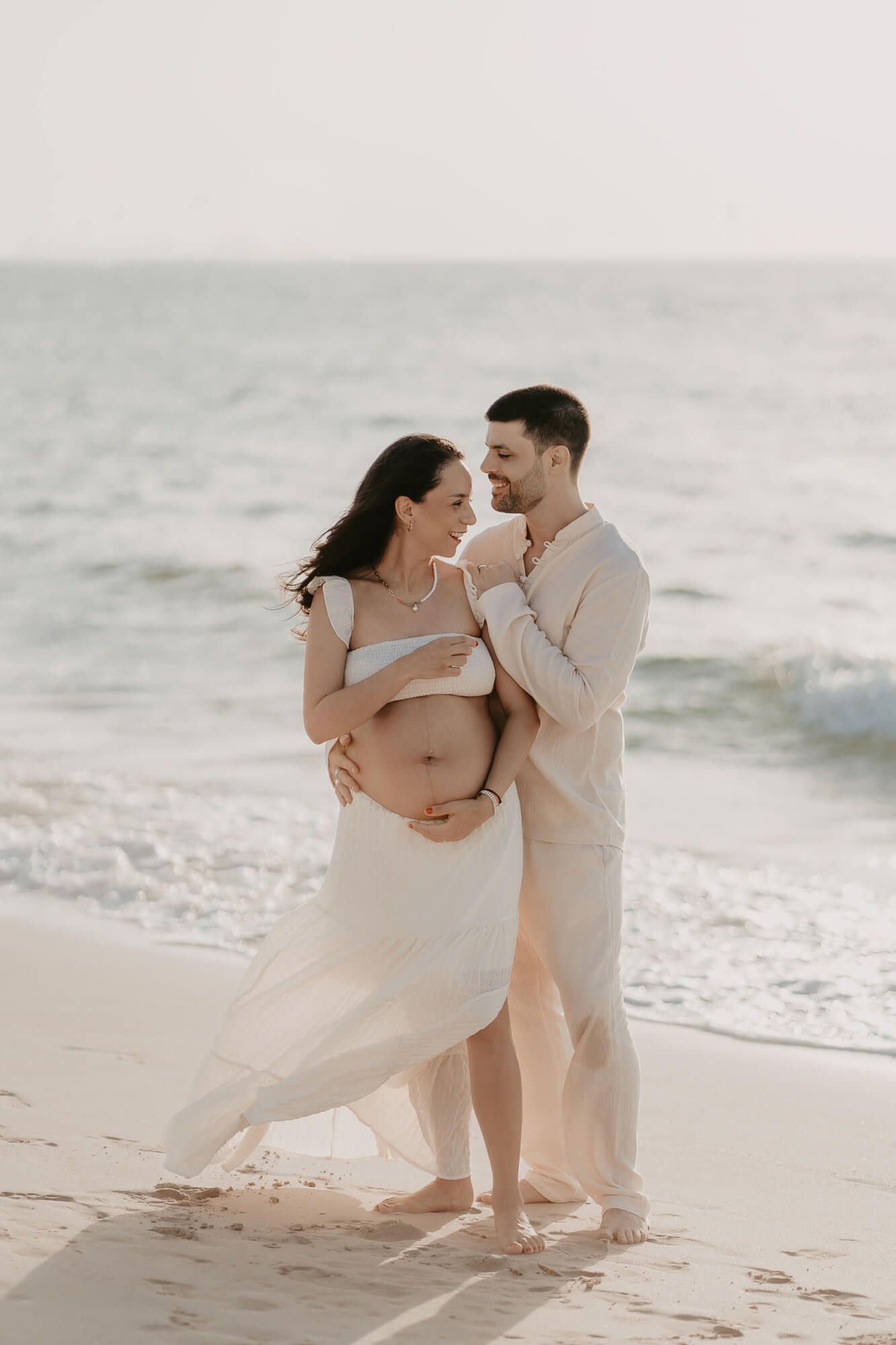 A couple in light beige clothing embraces on a misty beach during a maternity photoshoot.