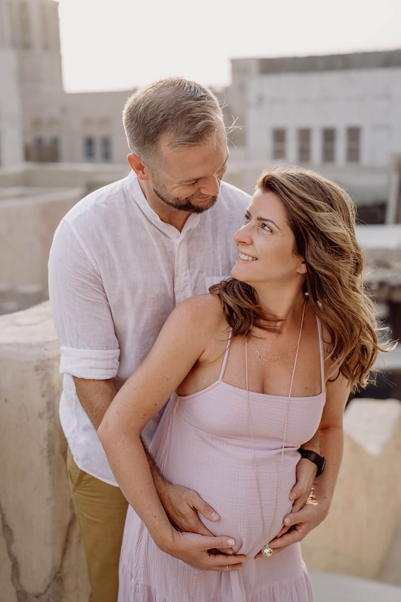 Rooftop maternity photo at sunset with couple in light pink and white outfits sharing intimate moment.