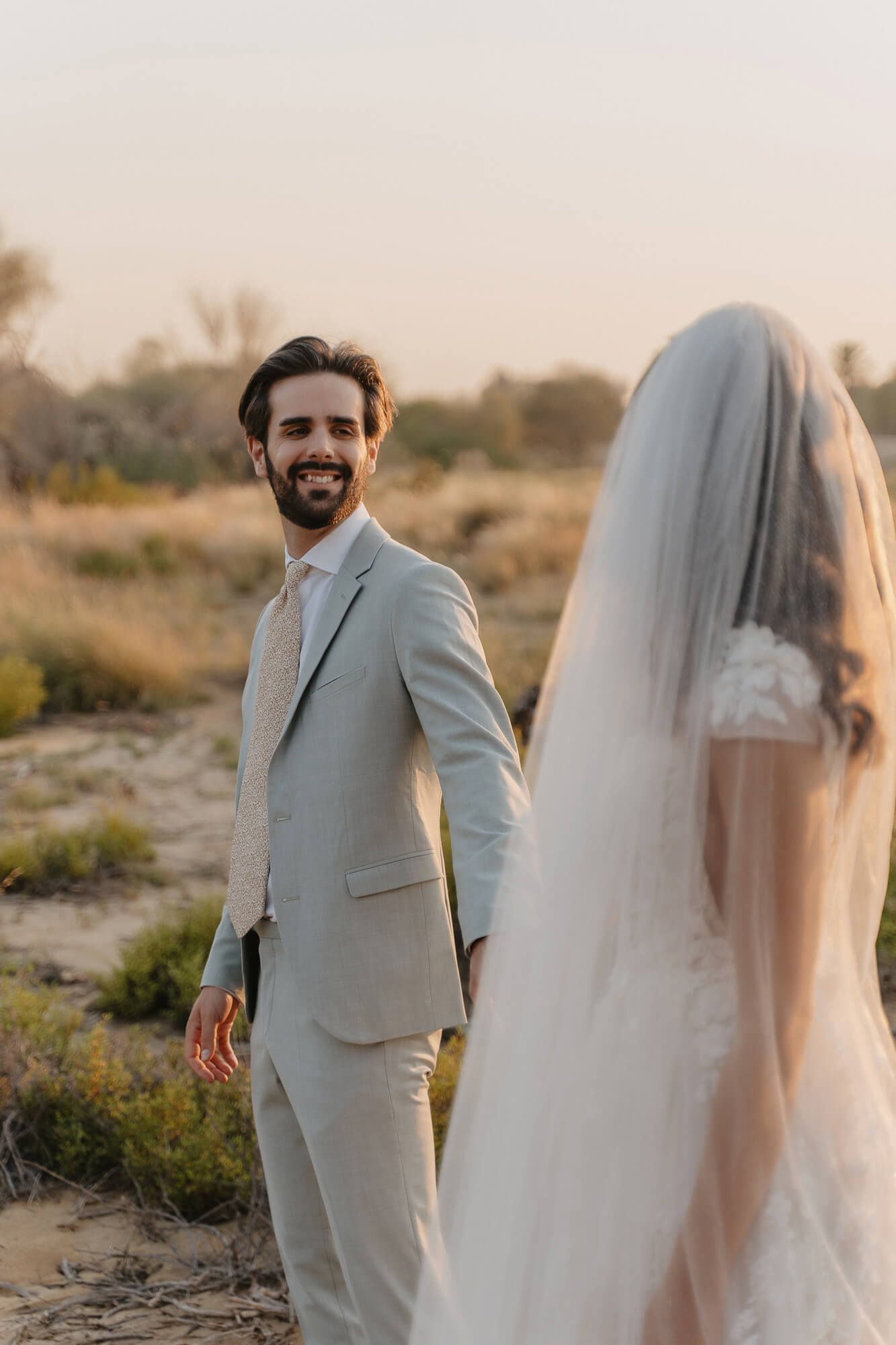 A couple stands in an outdoor desert setting at sunset for their wedding photos, with the bride in white and groom in gray suit.