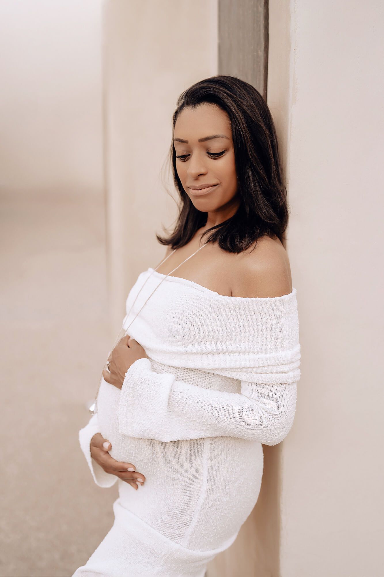 An expectant mother in a white dress poses with hands on her baby bump.