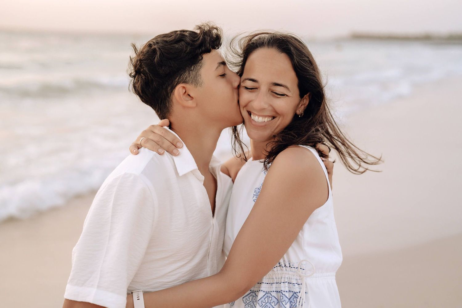 A couple shares an intimate moment on the beach, dressed in white as they smile and embrace on a sunny day.