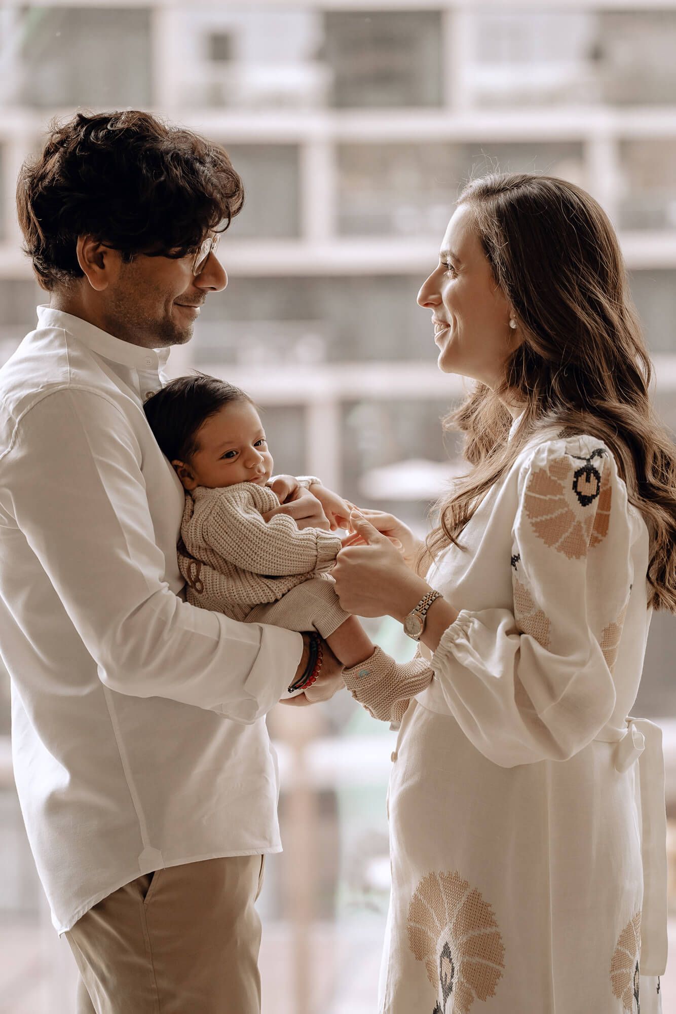 A tender family moment captured as parents smile while holding their baby near a window.