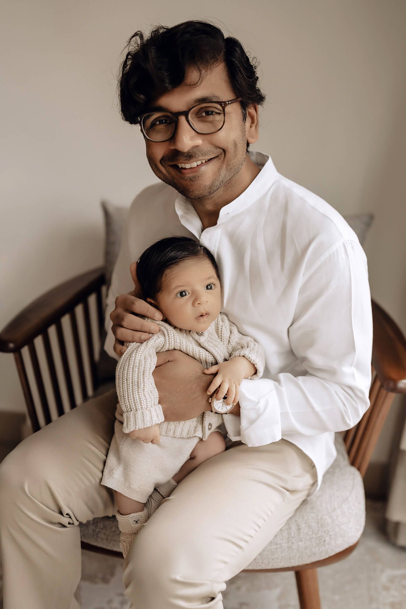 Parent and infant in coordinating outfits pose together on wooden chair.