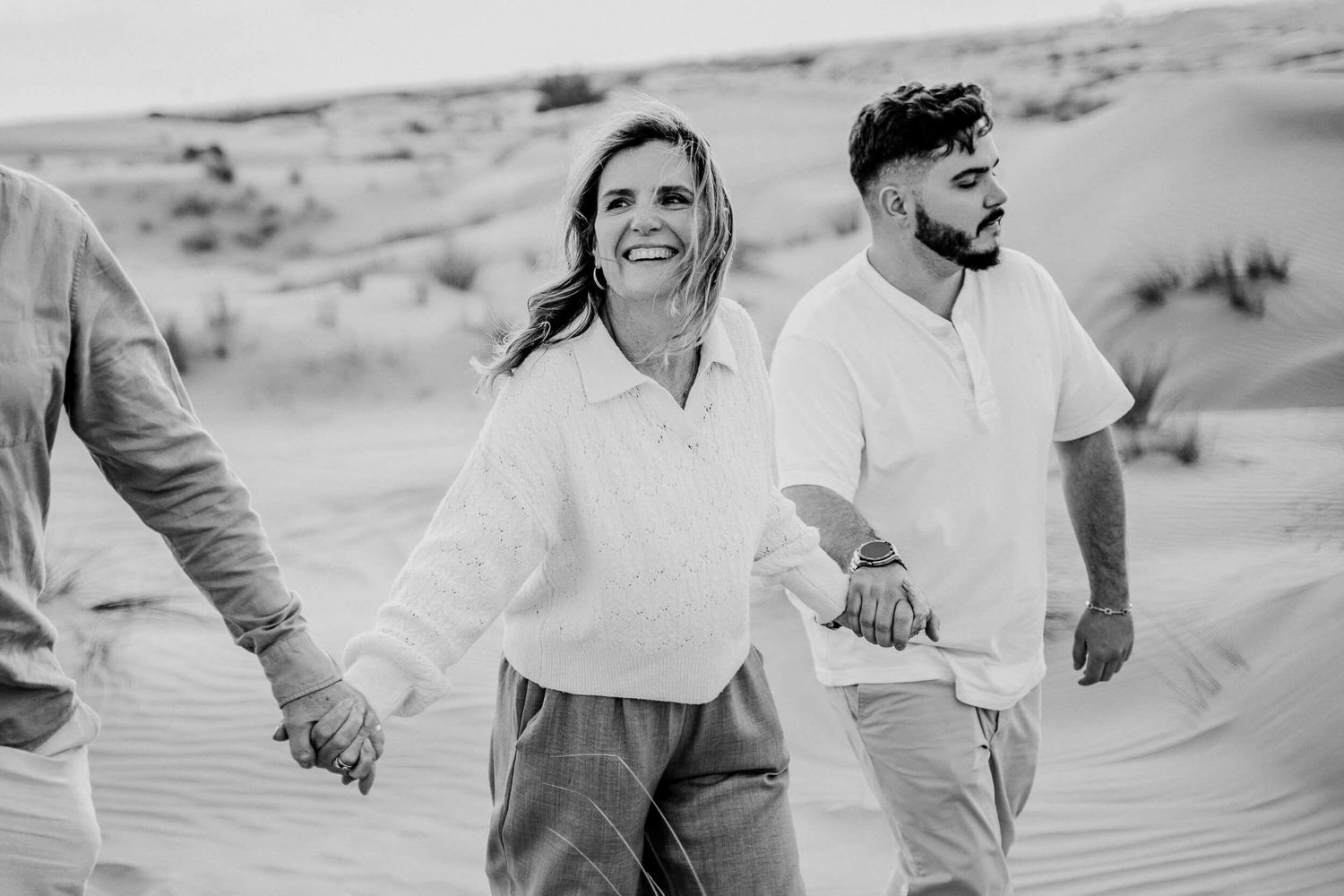 Black and white photo of people laughing while walking hand in hand through desert sand dunes.