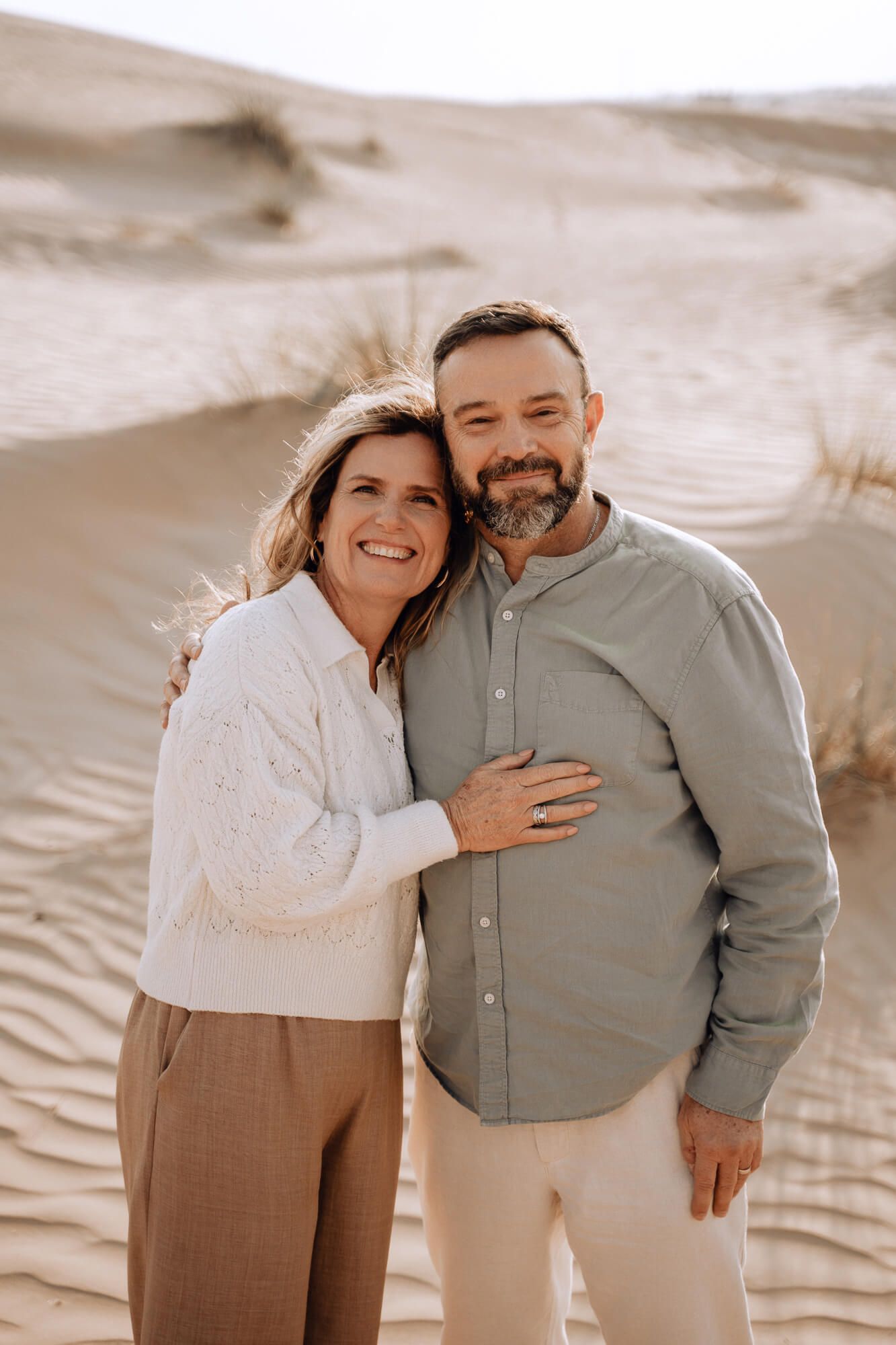 Couple smiling and embracing in front of desert sand dunes during golden hour.