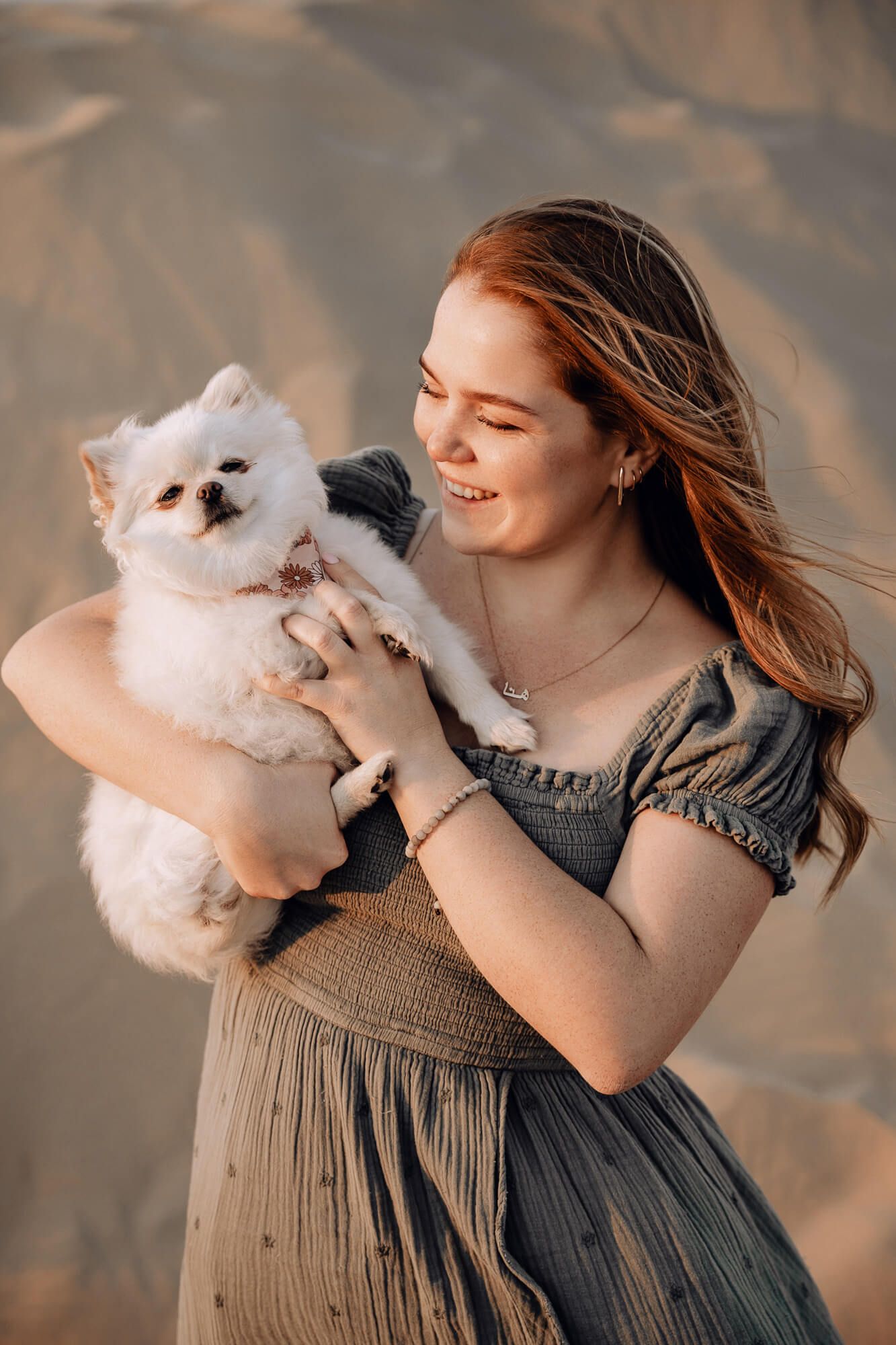Woman in gray dress holding fluffy white Pomeranian dog against desert backdrop at sunset.