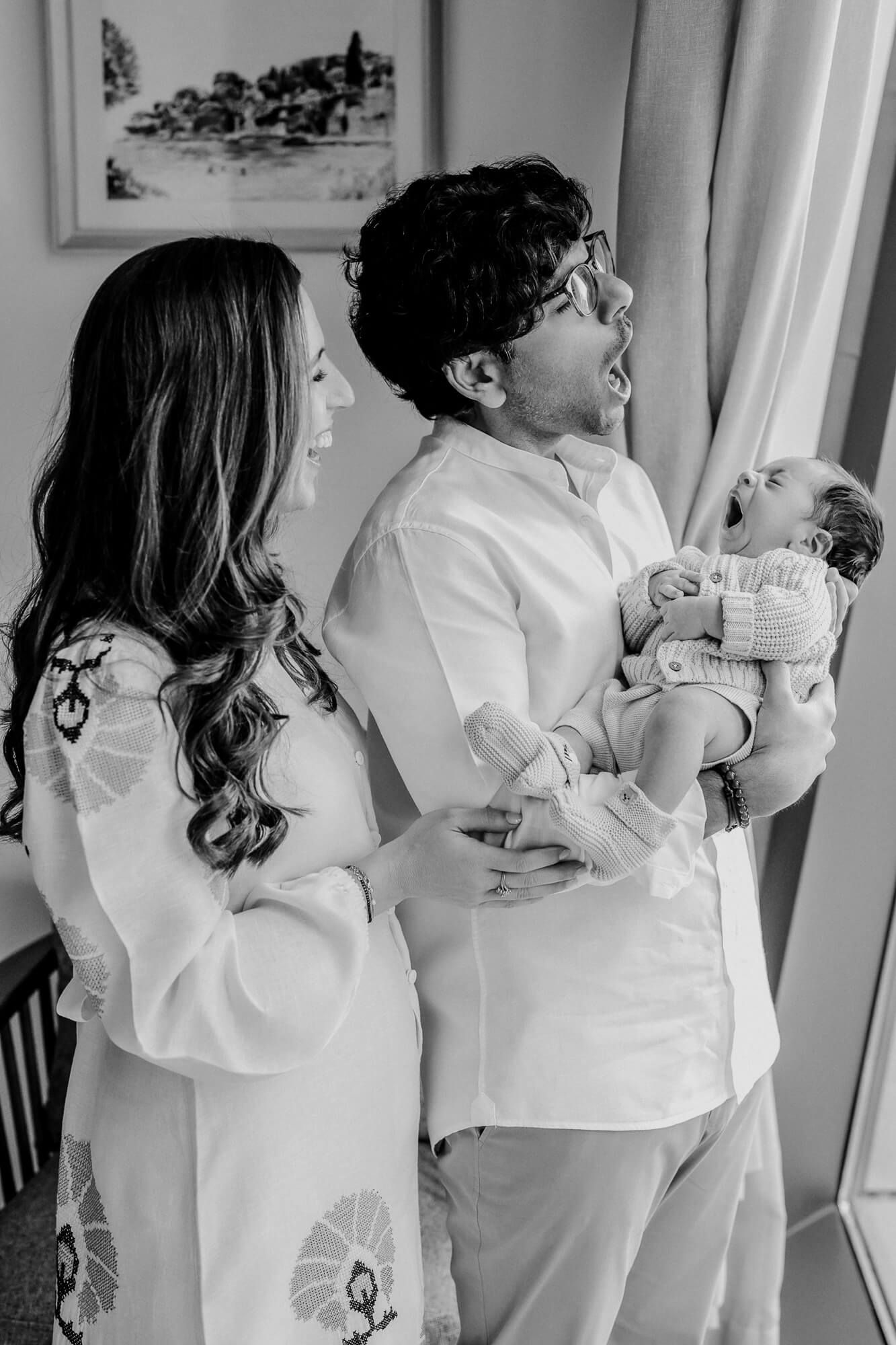 A black and white portrait of parents sharing a tender moment with their baby by a window.