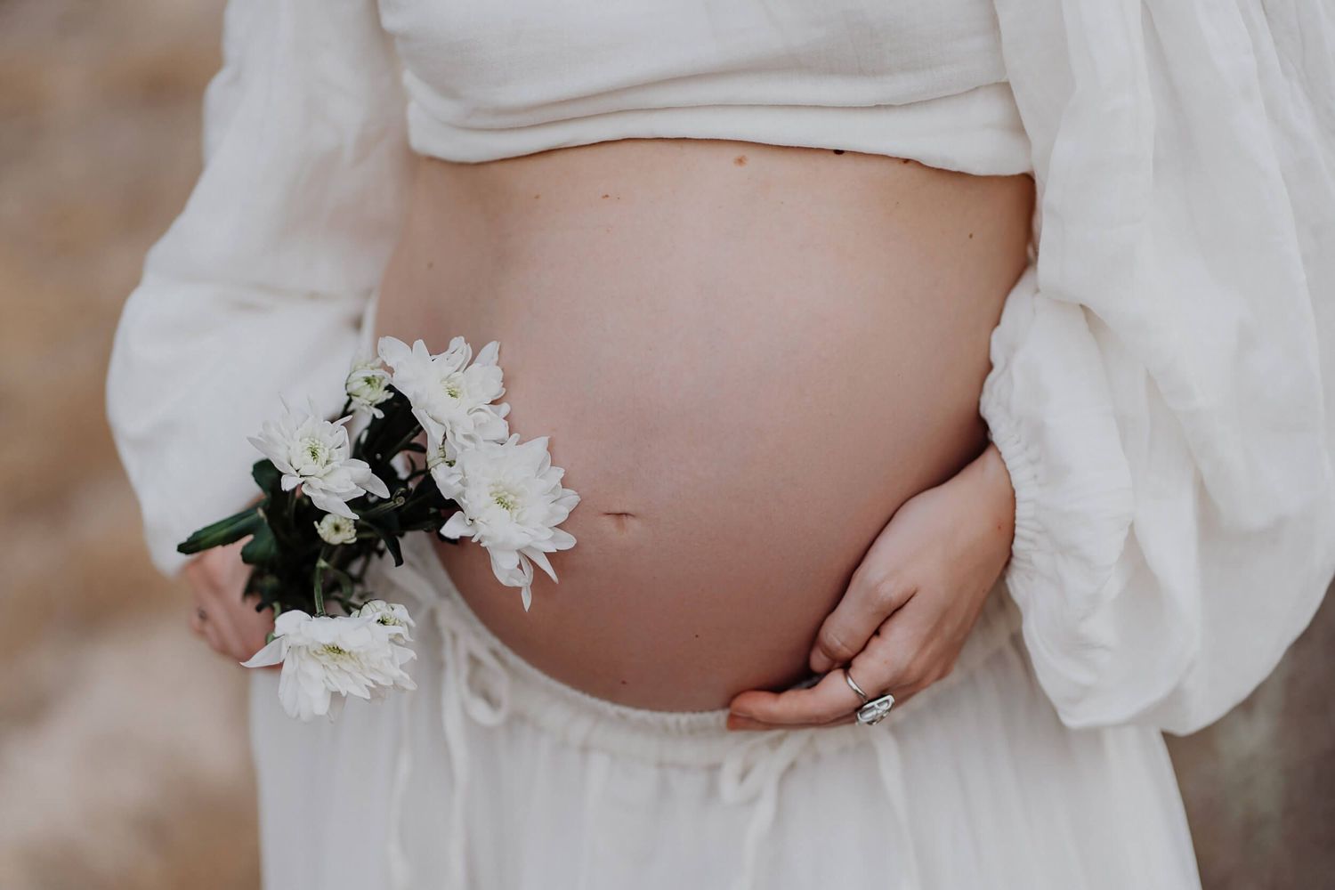 Pregnant belly in white dress holding white daisies against a soft neutral background.
