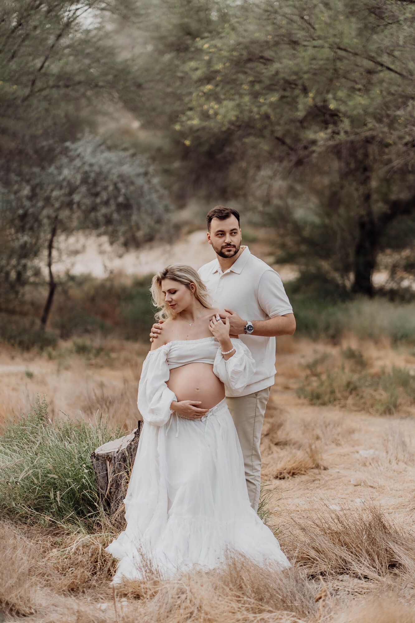 Desert maternity photoshoot featuring couple in dreamy white outfits among trees.