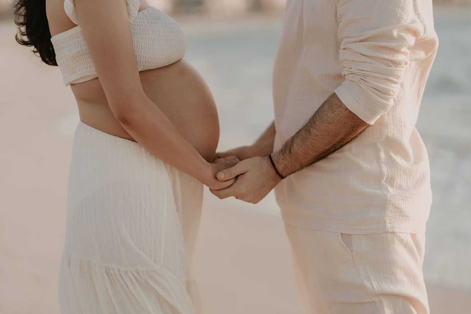 A couple in white outfits hold hands on the beach during a romantic photoshoot.