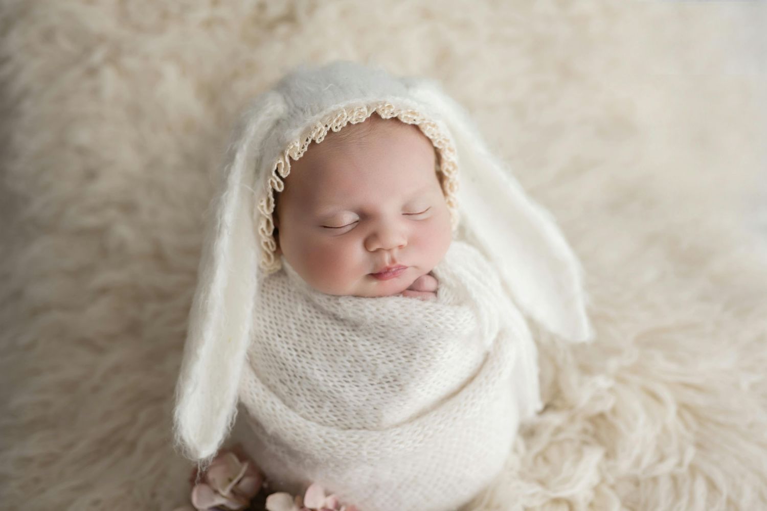 Sleeping newborn baby swaddled in soft white textiles on fluffy cream-colored blanket.