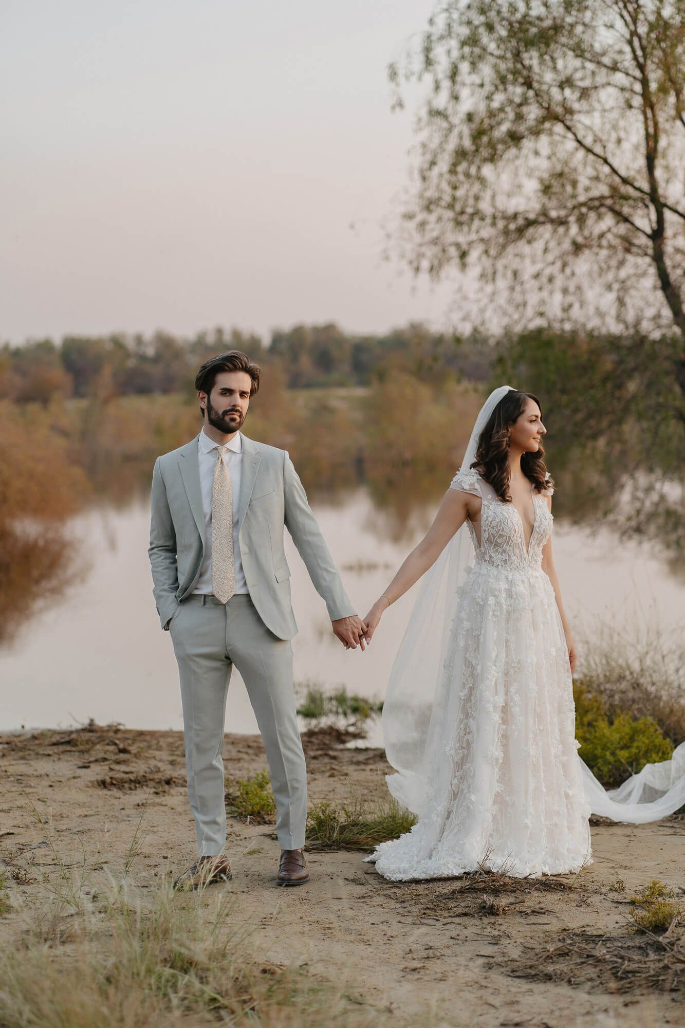 Wedding couple poses along a serene riverside landscape during autumn in matching grey and white attire.