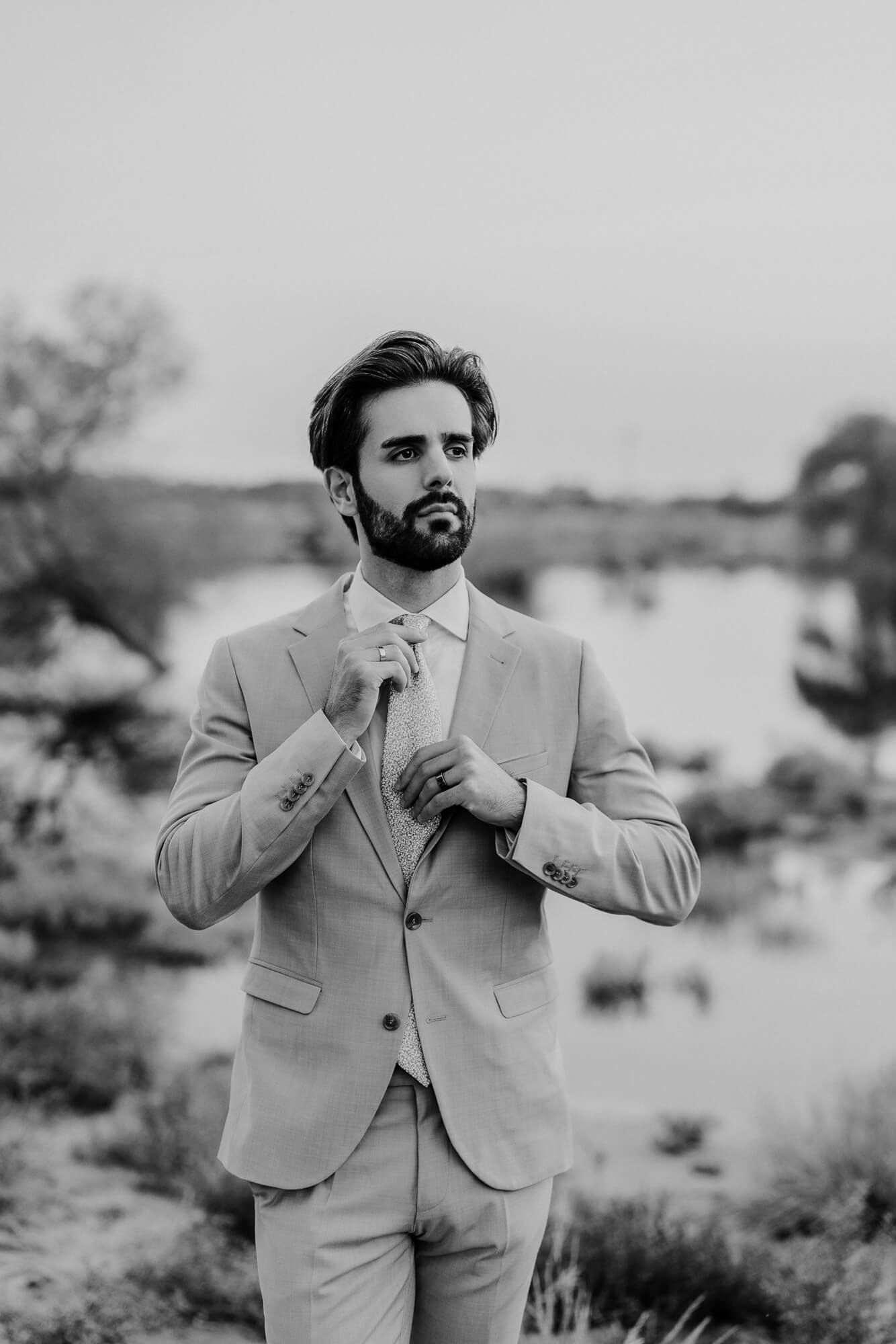 Professional black and white series showing a person in a light-colored suit adjusting their tie outdoors in nature.