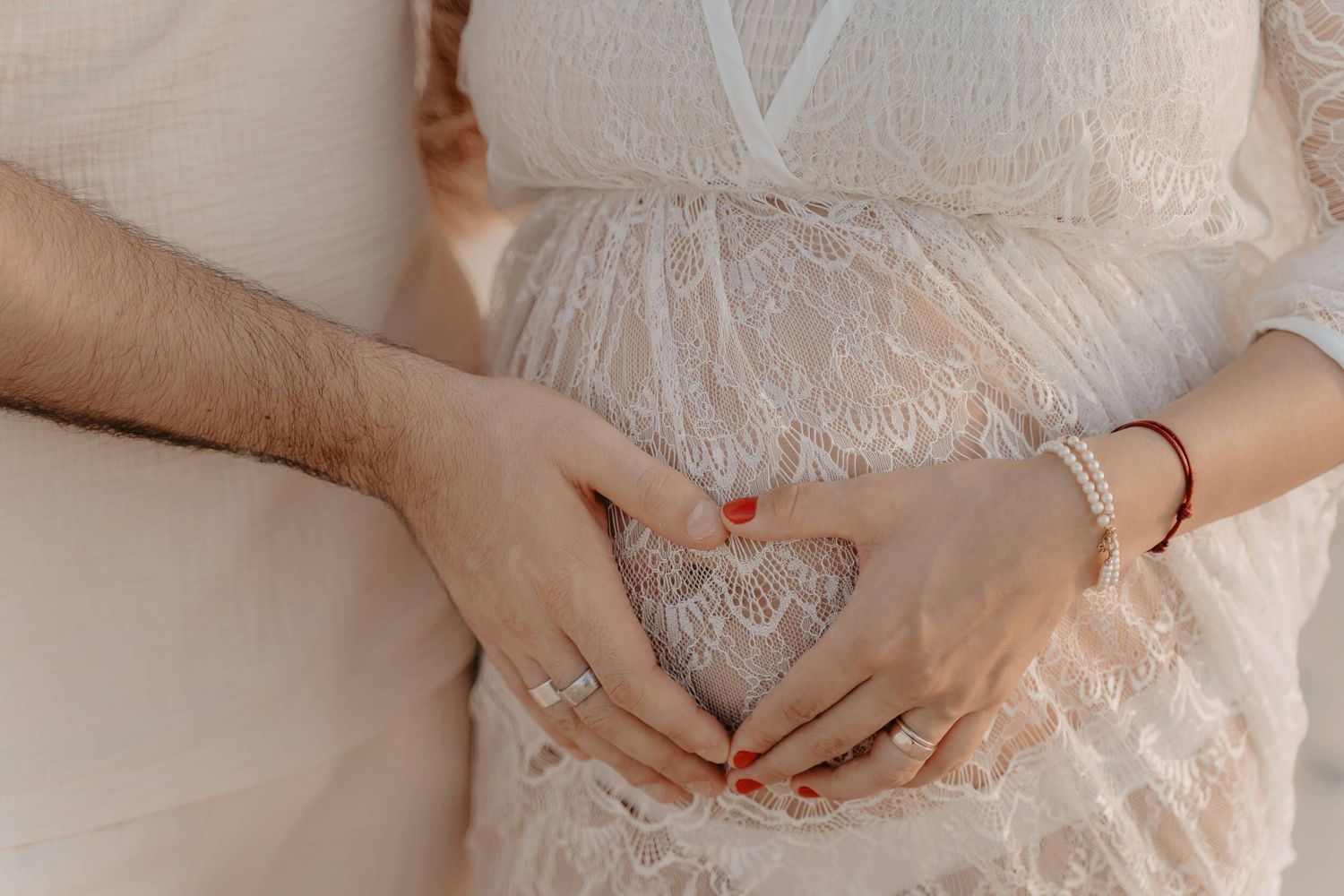 Hands forming heart shape over pregnant belly wearing delicate white lace maternity dress.