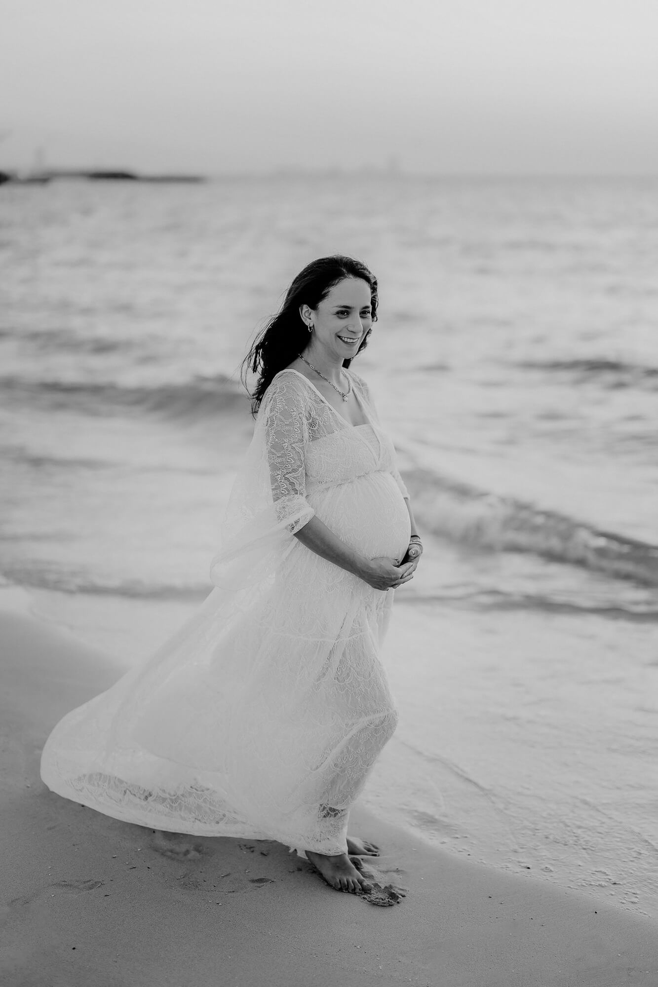 Black and white photo of an expectant mother in a flowing lace dress walking along a beach at sunset.