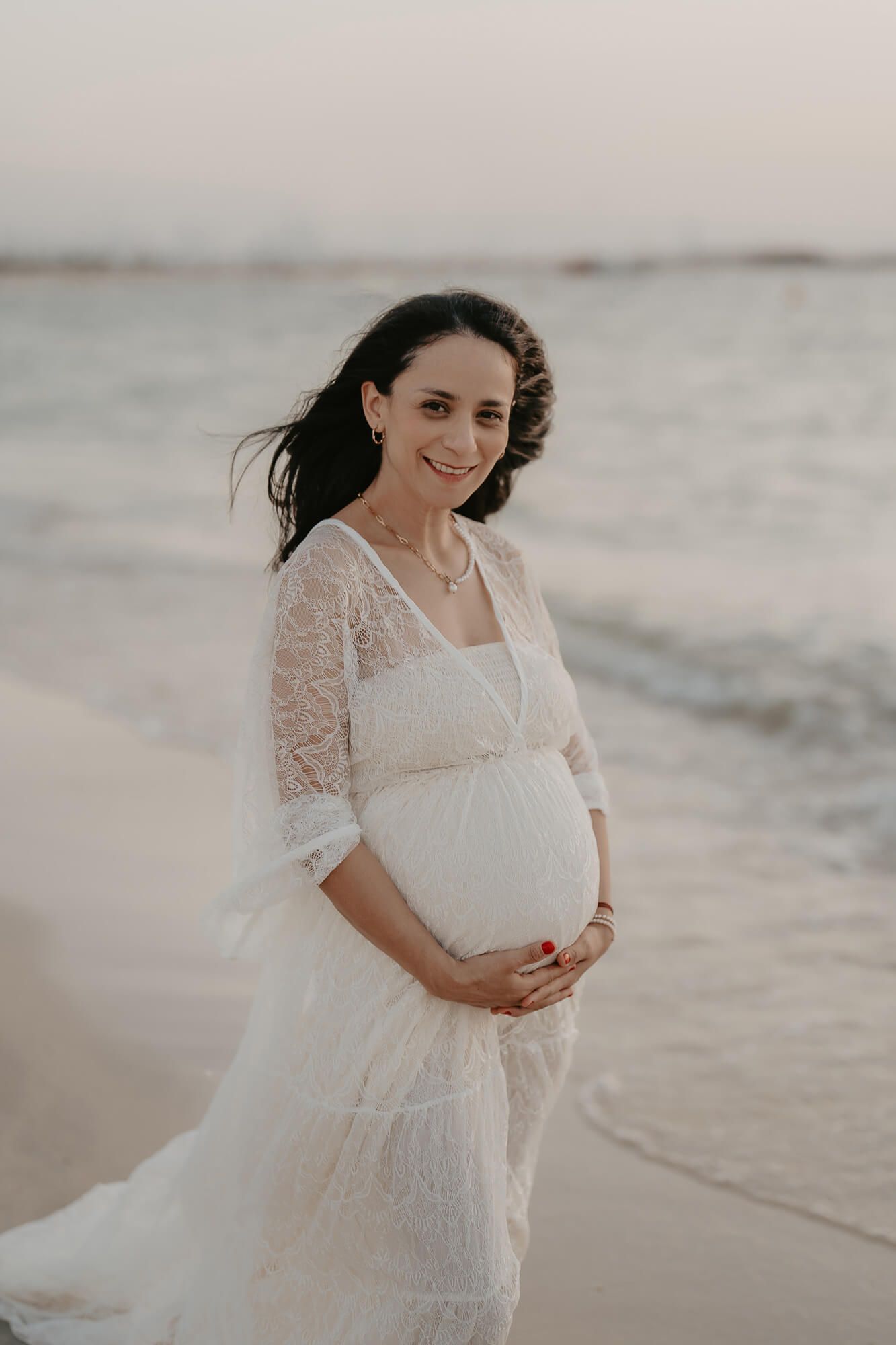 Expectant mother in flowing white lace dress poses on beach at sunset with wind in her hair.