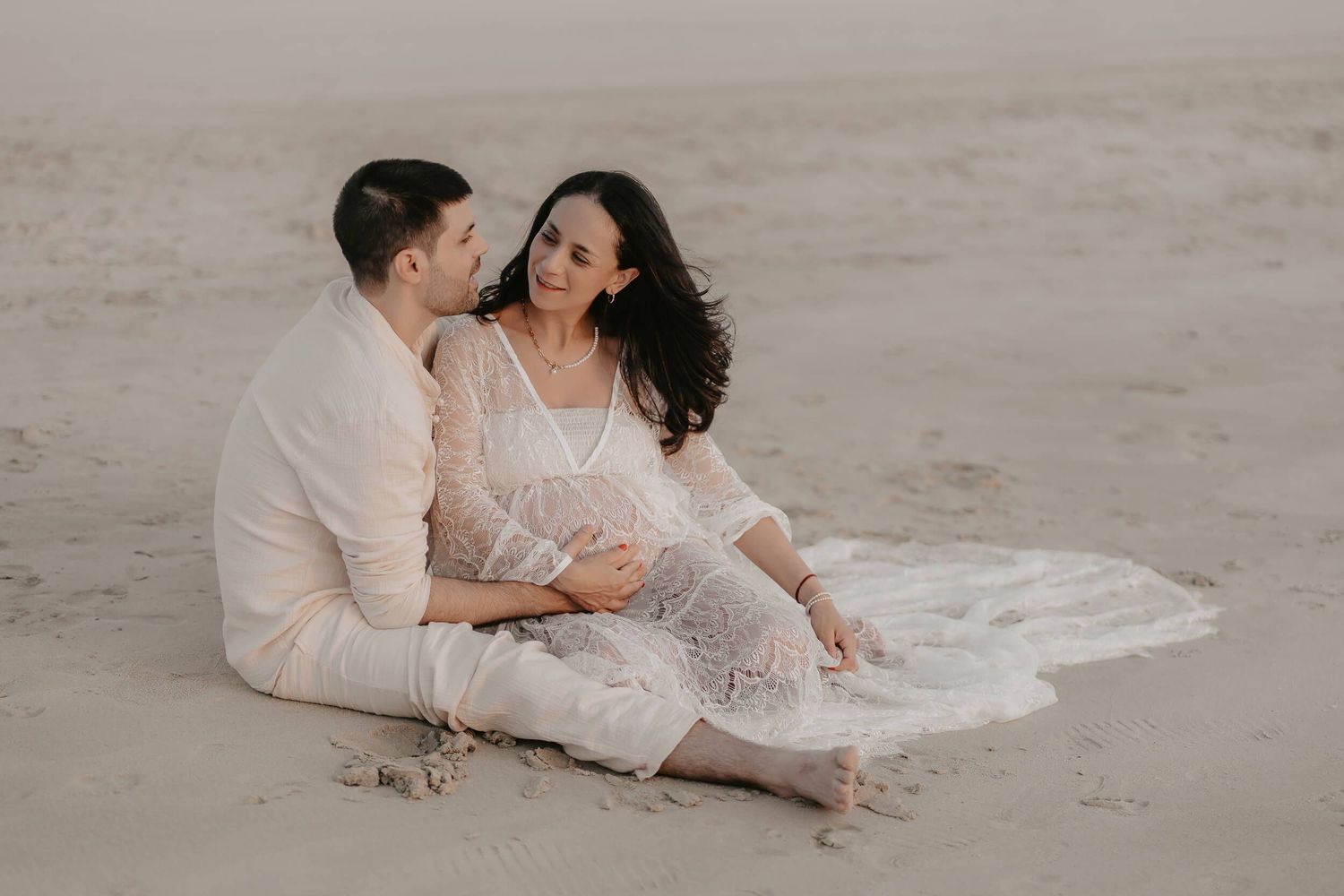 A couple in white clothing sits together intimately on a sandy beach during sunset.