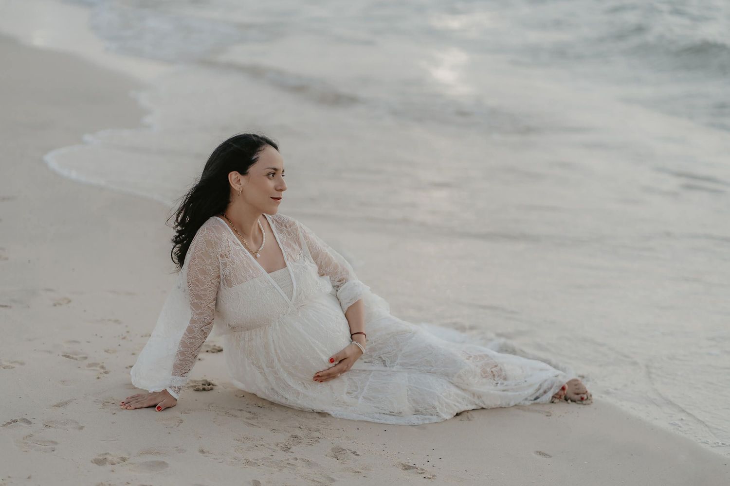 Elegant figure in flowing white dress reclining on sandy beach at sunset near ocean waves.