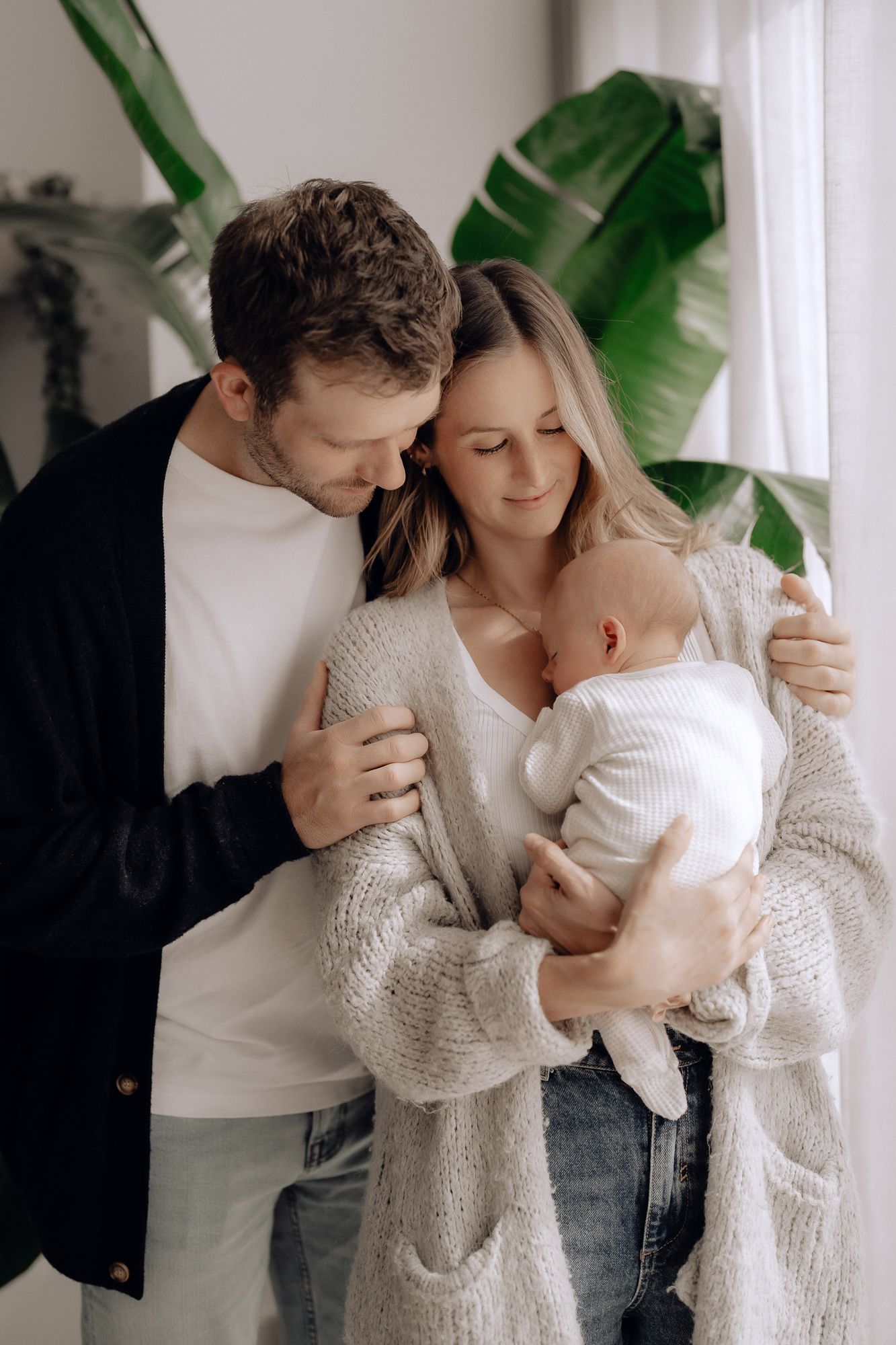 A tender family portrait with parents embracing their baby against a backdrop of tropical green leaves.