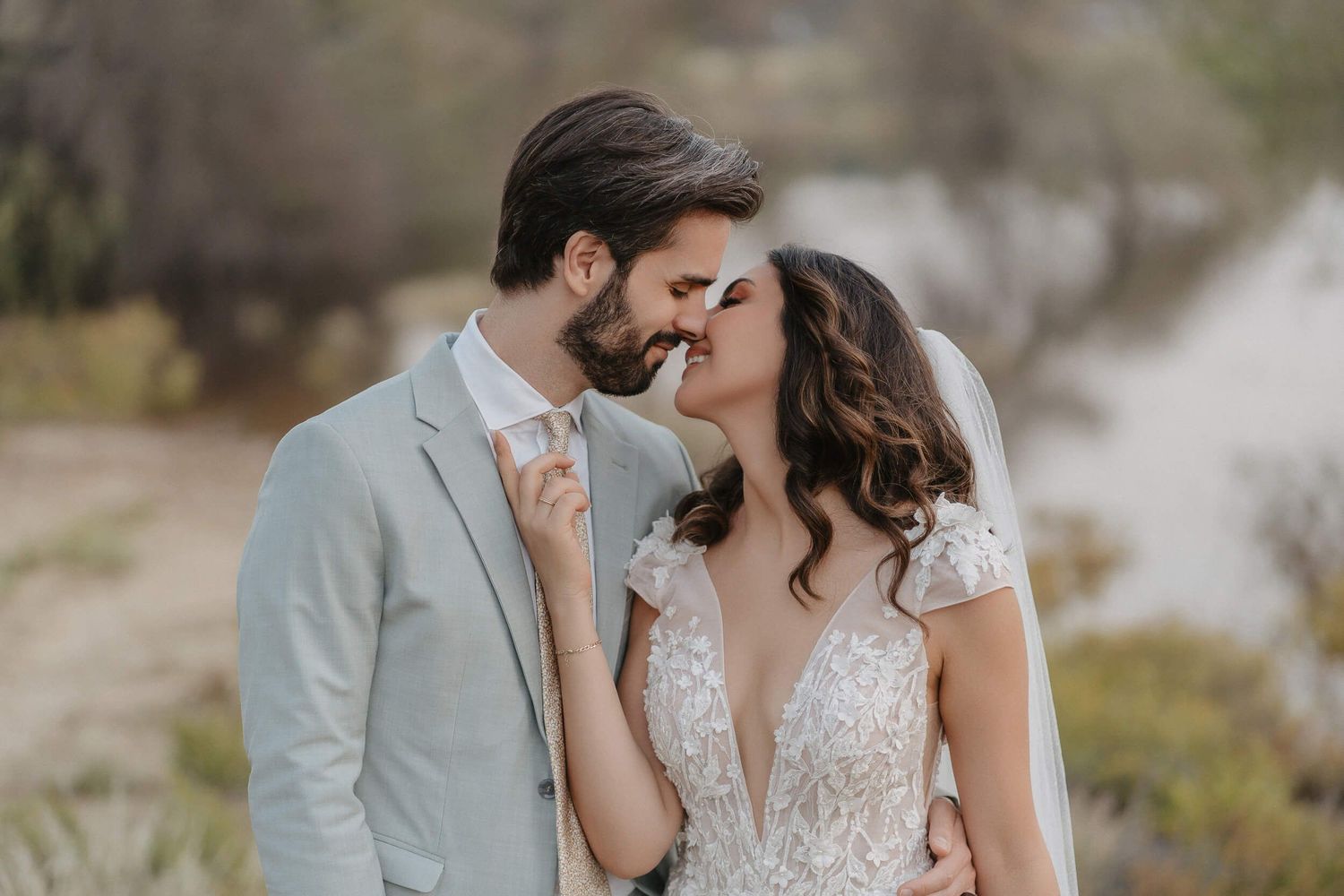 Romantic bridal couple shares intimate moment outdoors in neutral tones with lace wedding gown and light suit.