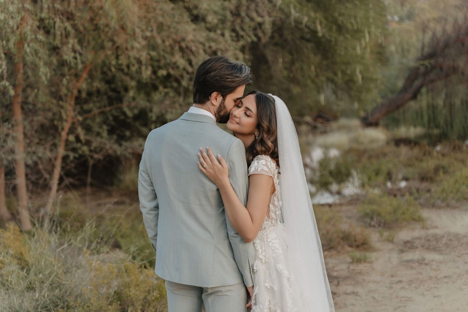 A romantic wedding couple embraces on a rustic desert path surrounded by natural brush and soft lighting.