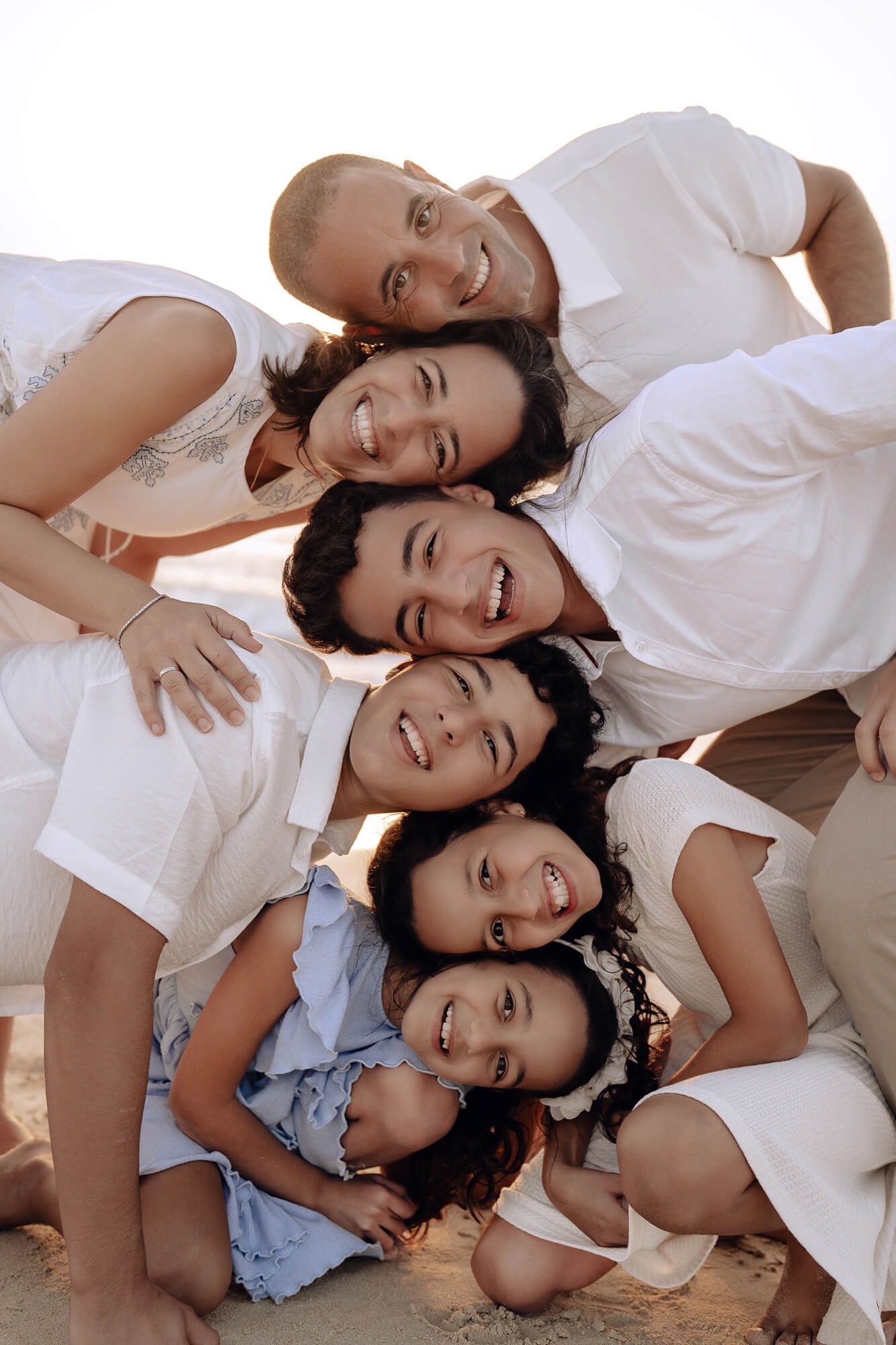 A joyful family in white clothing lies together on the floor while posing playfully for a casual photo shoot.