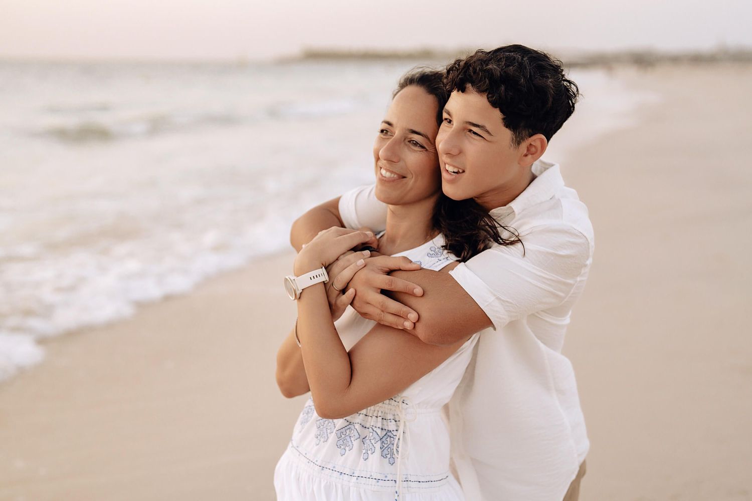 A romantic couple embracing on the beach in white outfits against an ocean backdrop.