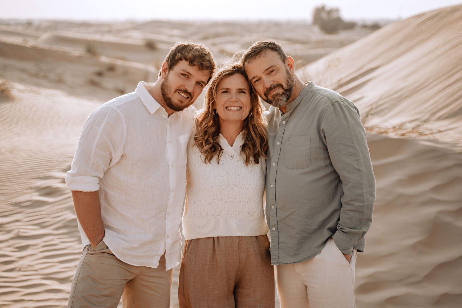 Friends share an intimate moment together while sitting on sand dunes wearing earth-toned clothing.