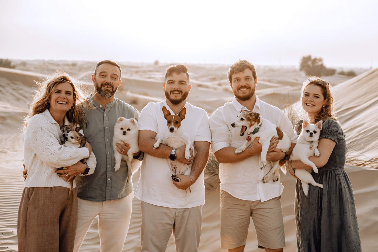 A casual group portrait on sand dunes with everyone holding small dogs and wearing light colored clothing.