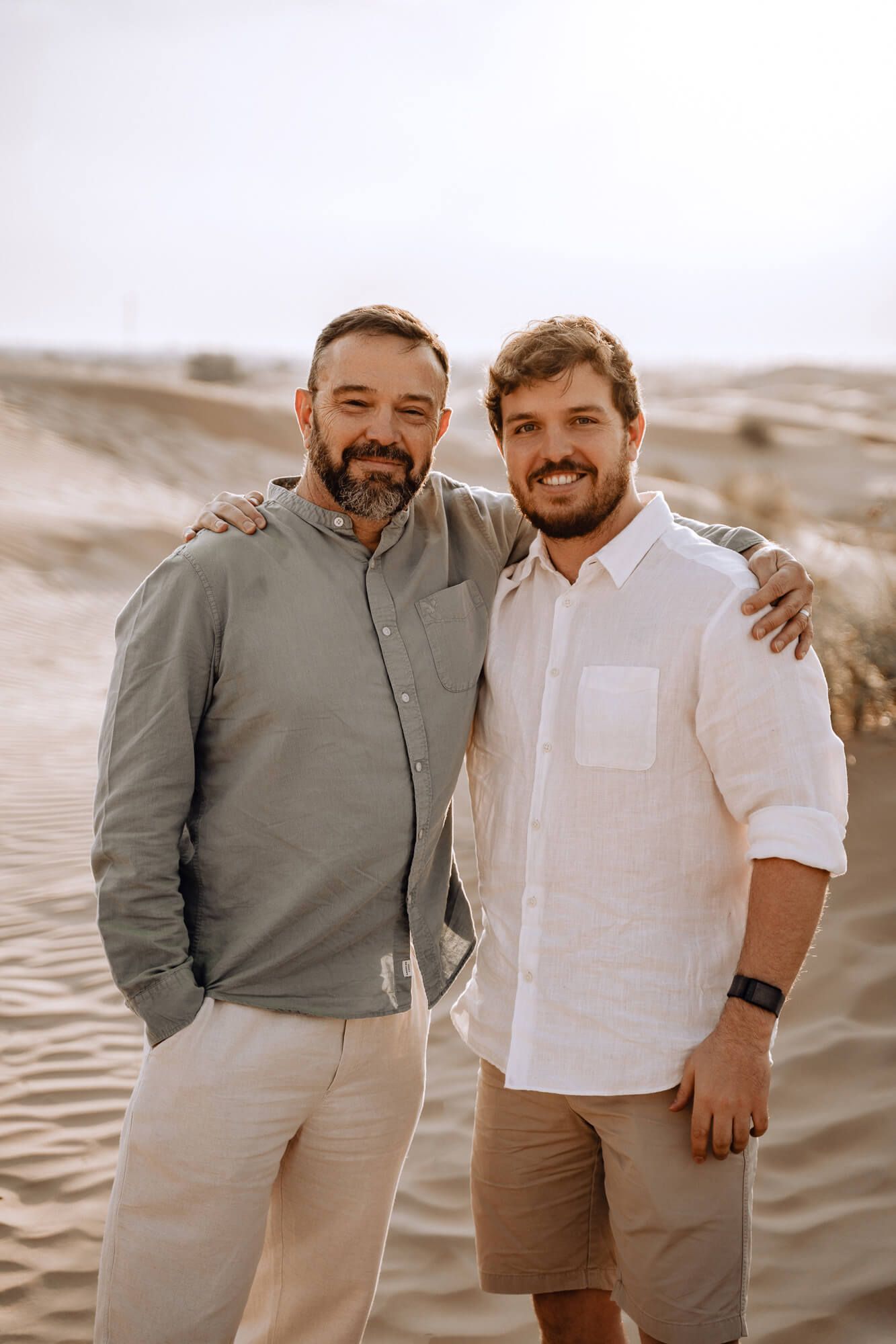 Two people pose closely together wearing coordinating casual outfits in sandy desert terrain.
