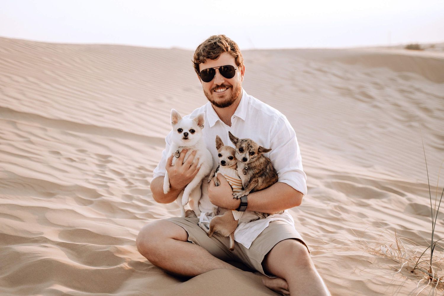 Person sitting on sand dunes holding two small dogs in a casual outdoor photo shoot.