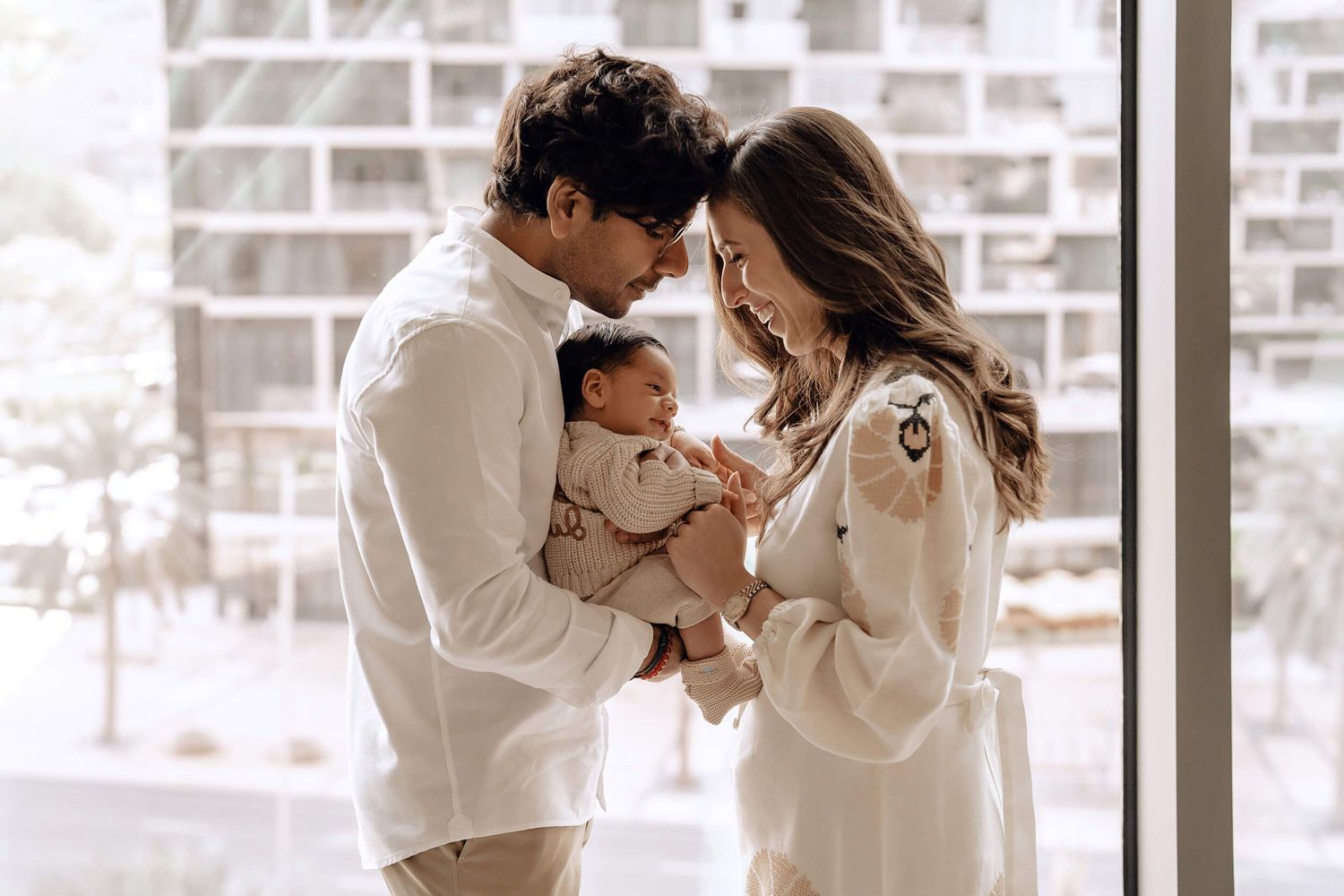 A couple in white outfits sharing an intimate moment with their baby by a bright window.