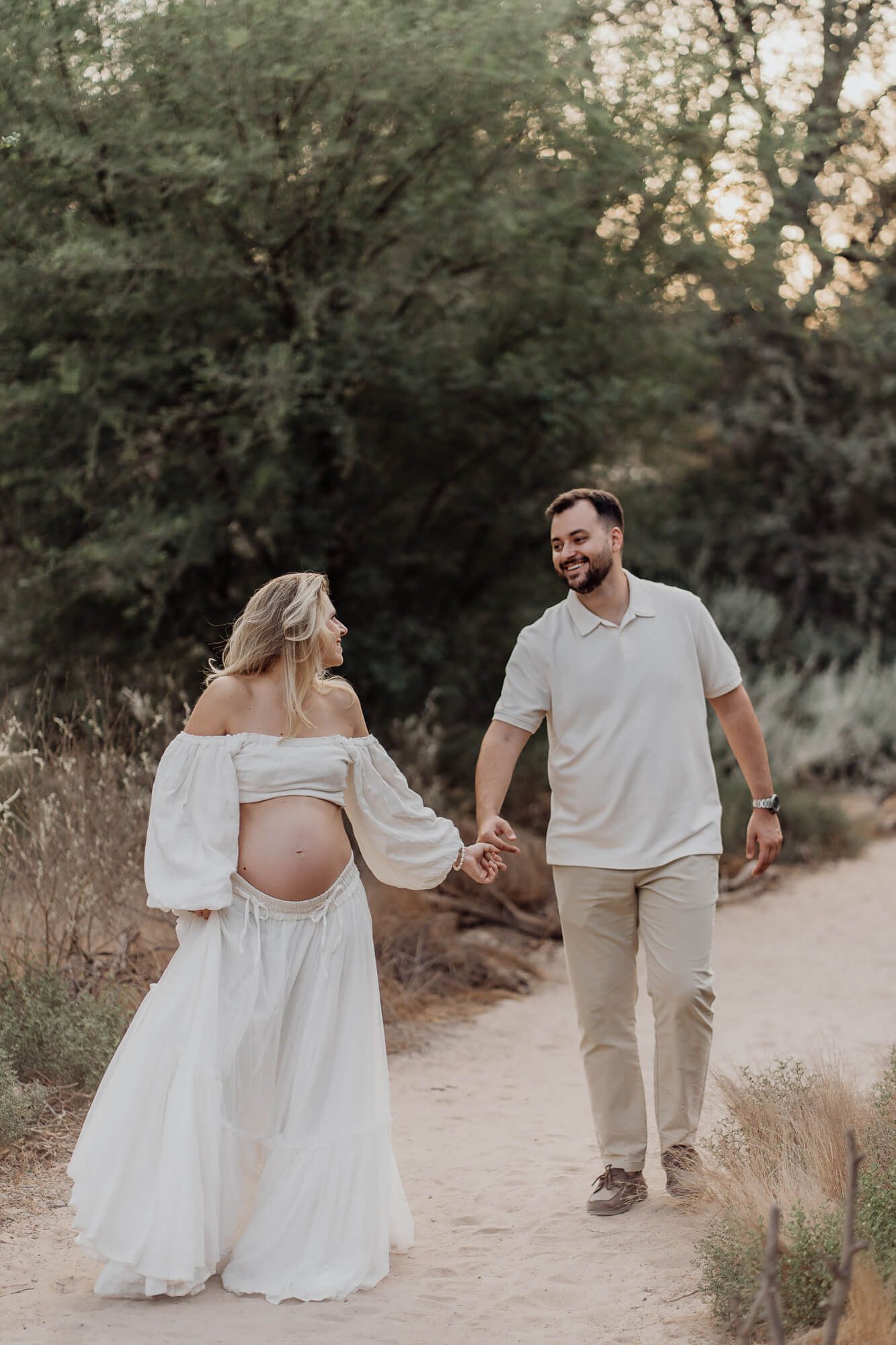 Maternity portrait of couple in coordinated outfits walking along natural path at dusk.