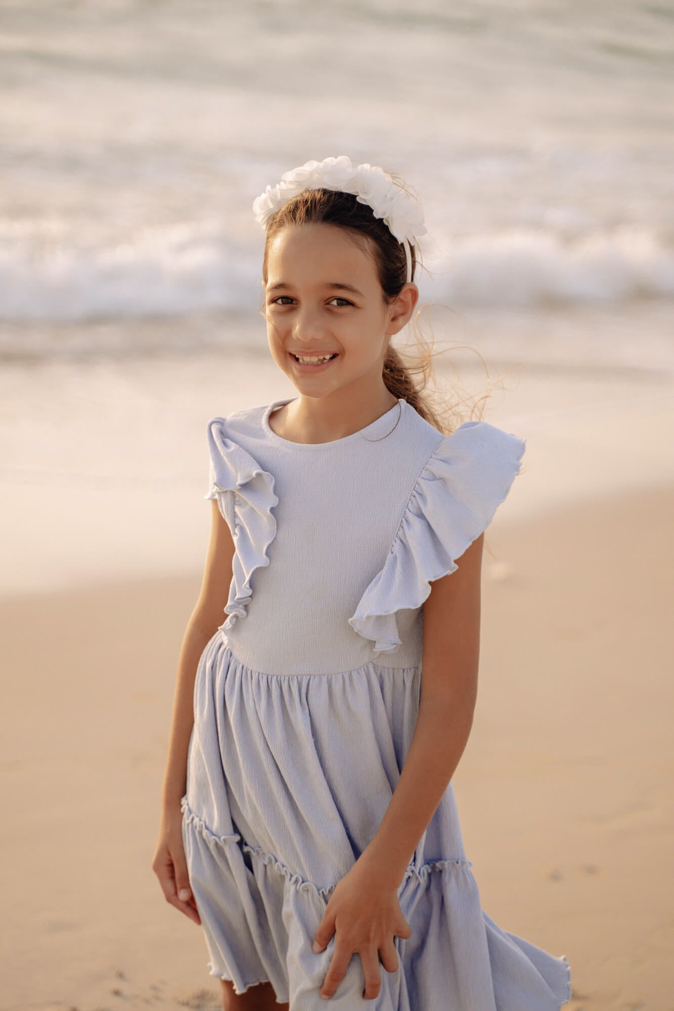 Young child wearing a light blue ruffled dress plays and smiles on a sandy beach at sunset.