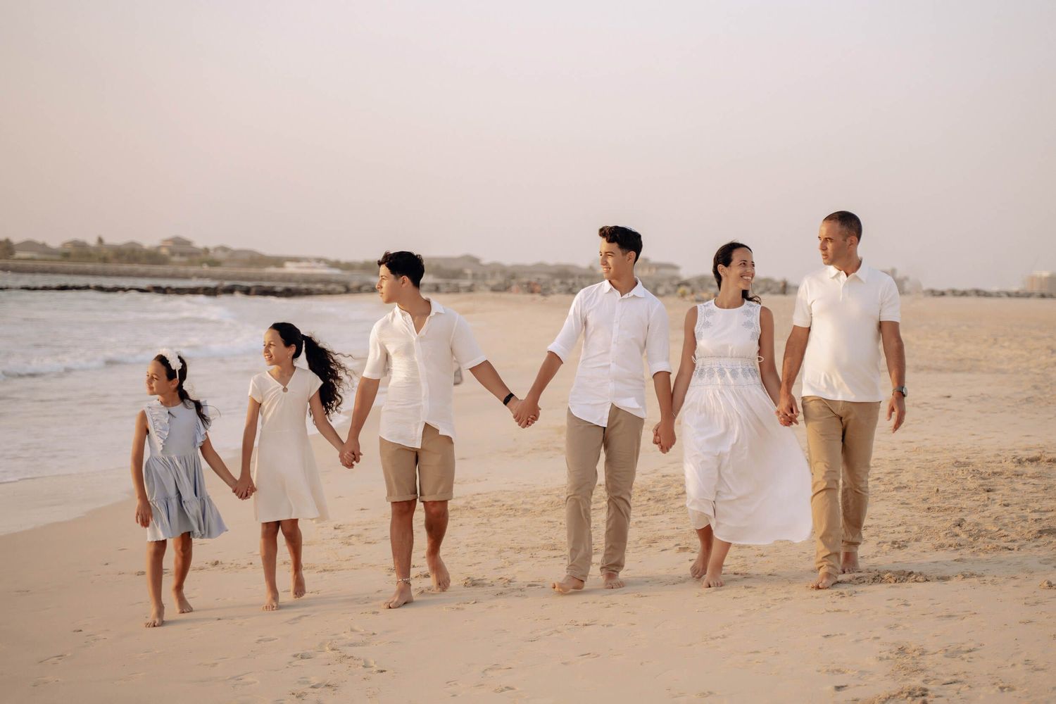 Family walking together hand in hand along a beach at sunset wearing coordinated white and beige outfits.