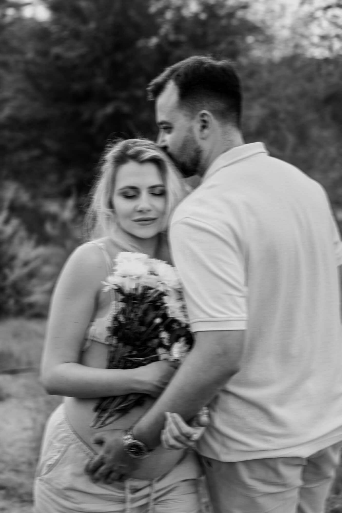 A couple shares an intimate moment while holding white flowers in a romantic outdoor setting.