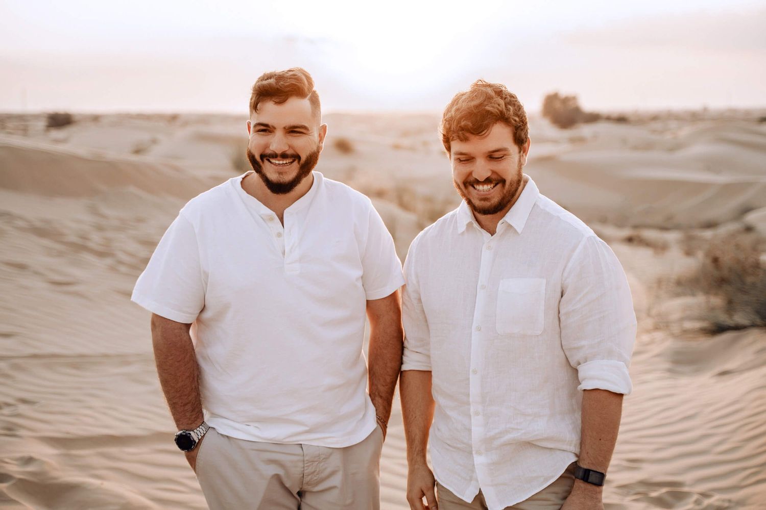 Two people in white shirts and khaki pants stand together on desert sand dunes.