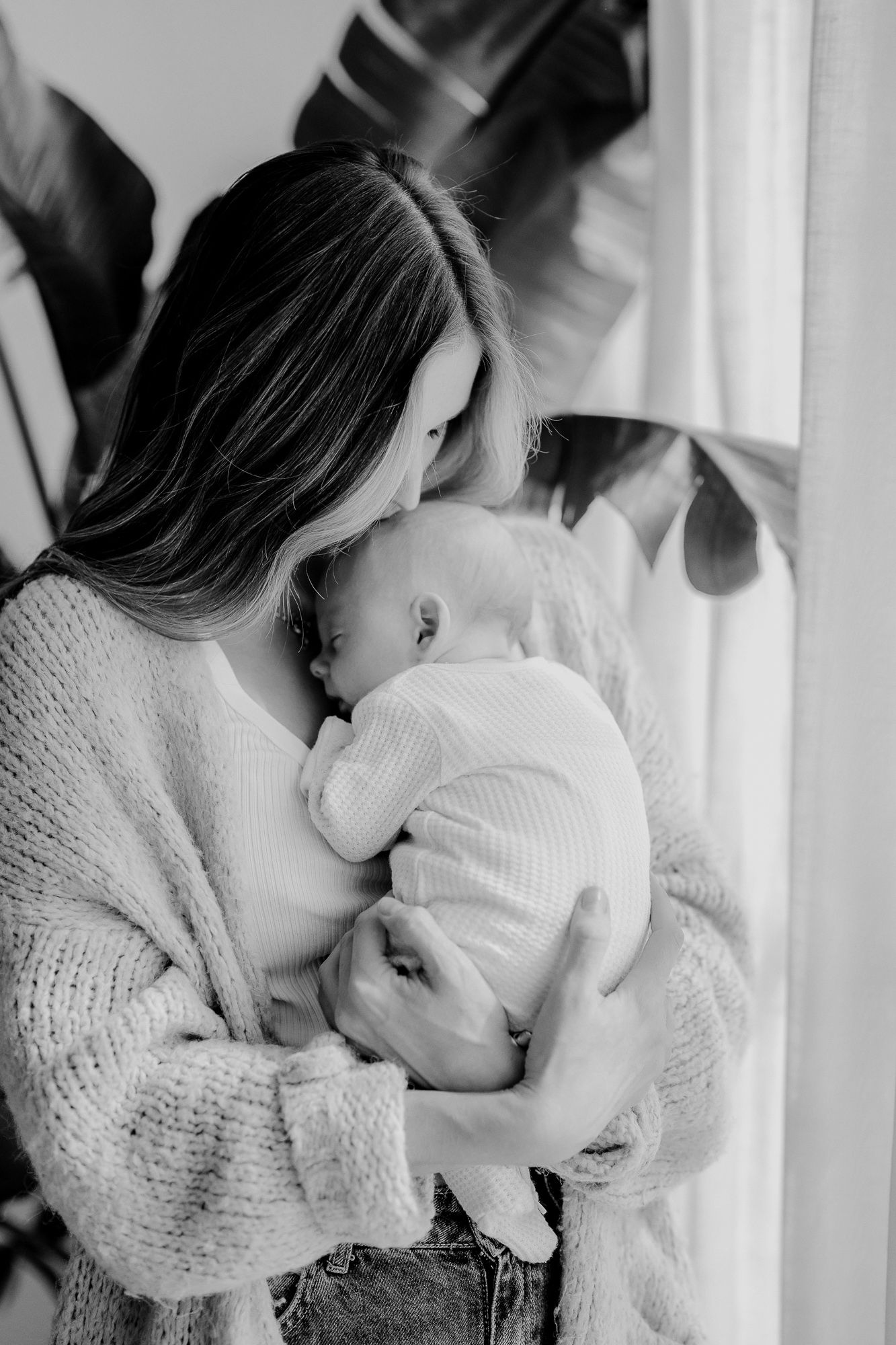 Tender black and white portrait of a mother in a cozy sweater holding and comforting her baby near a window.