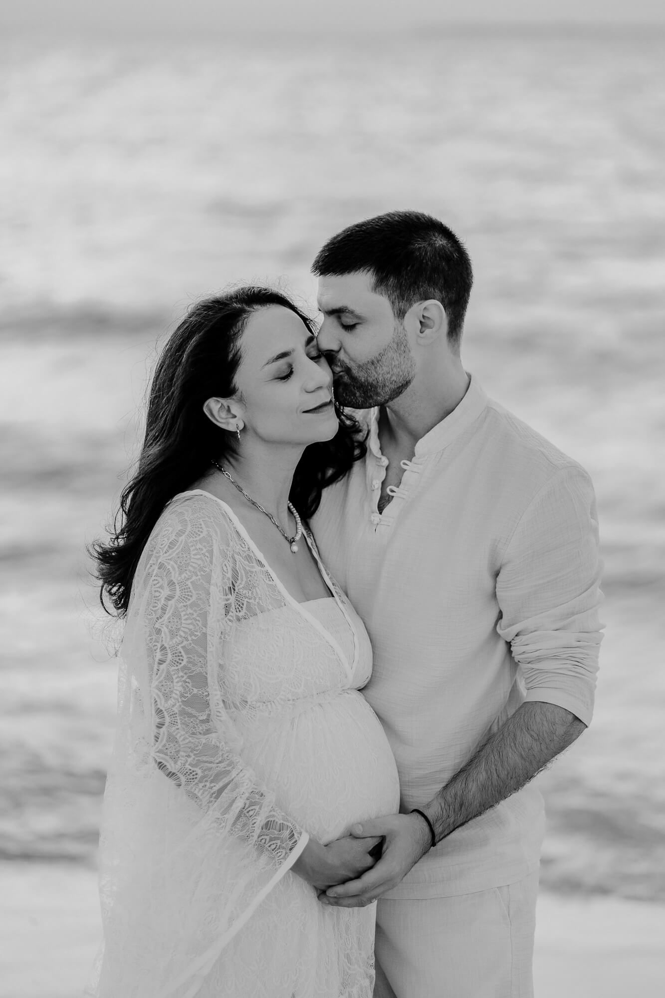 Black and white photo of an intimate beachside maternity shoot with couple embracing in white attire.