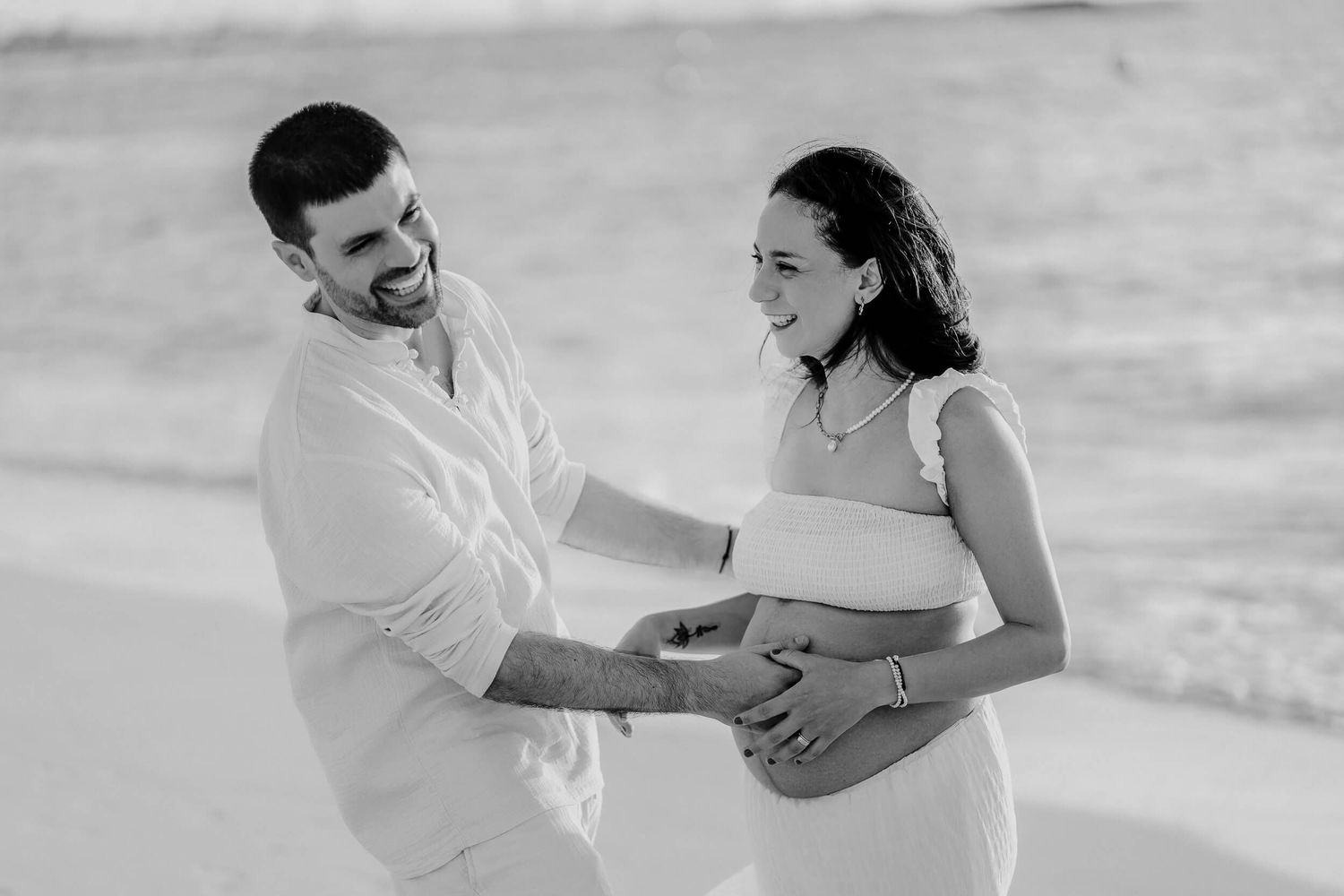 Black and white photo of a couple sharing joyful laughter while embracing on a sandy beach.