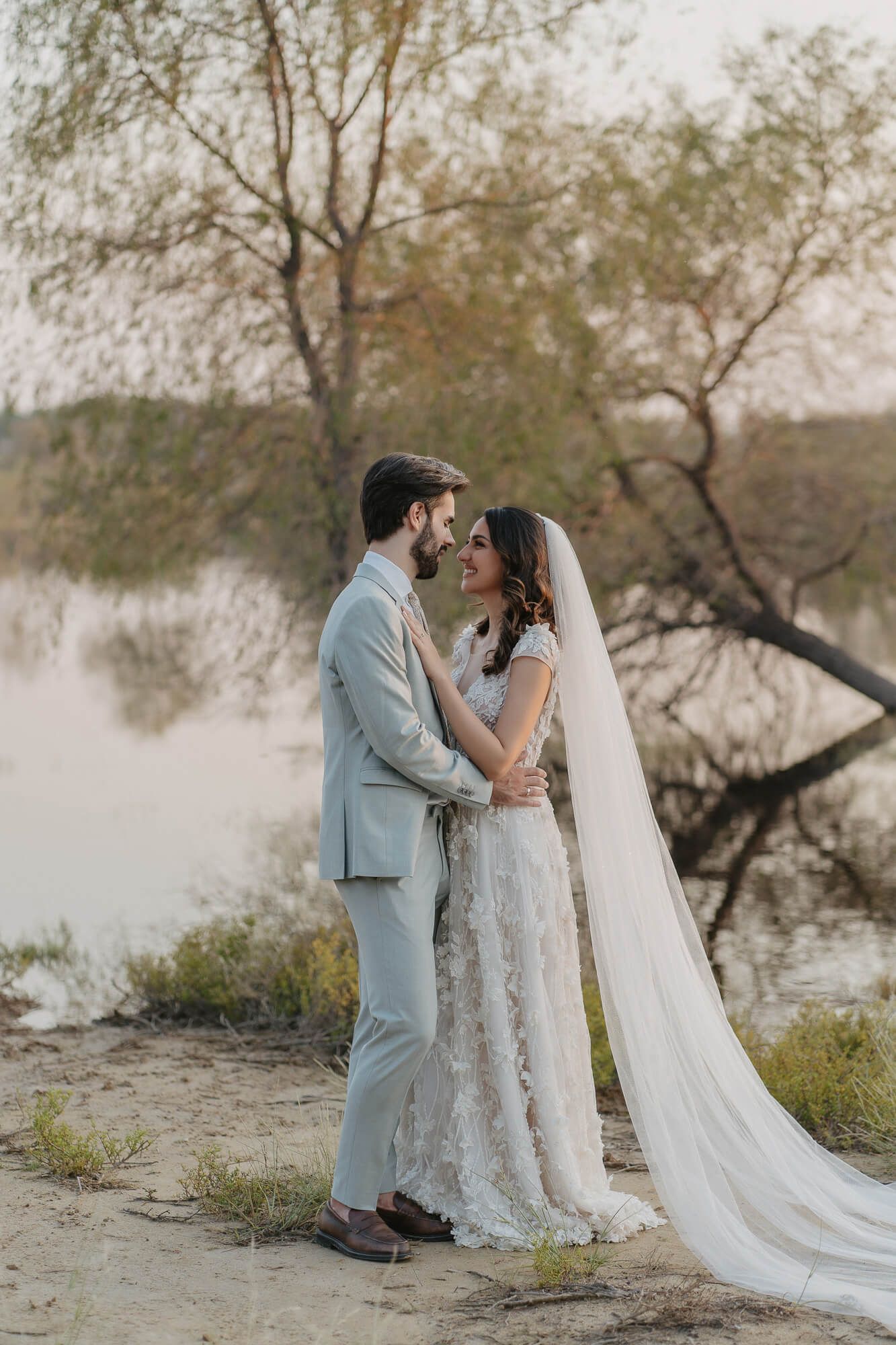 A romantic wedding couple embraces by a tranquil lake at sunset, with willow trees and a long flowing veil in view.