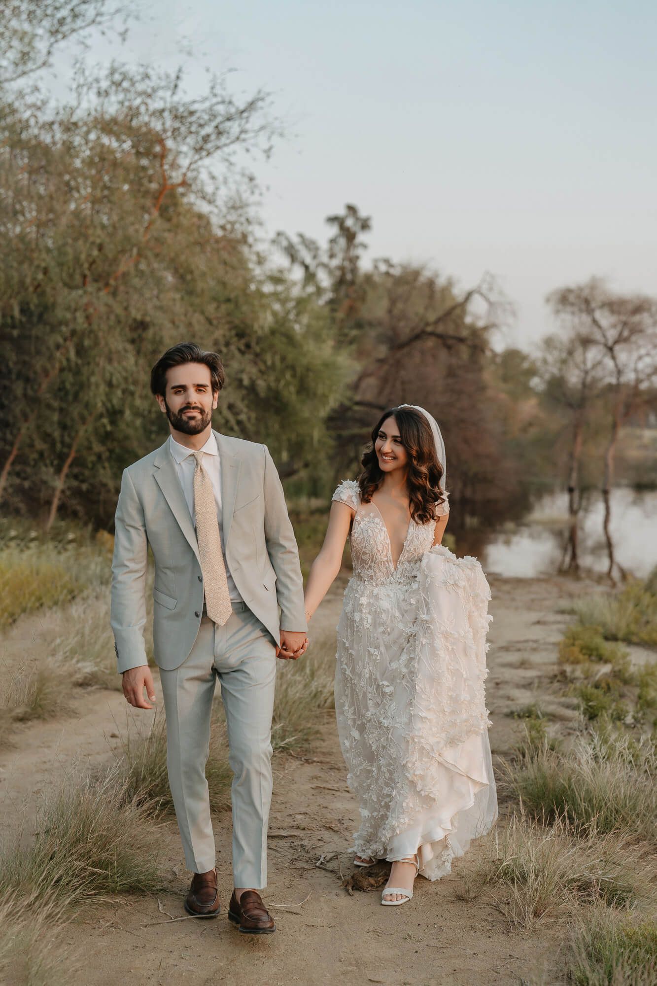 A couple in elegant wedding attire walking hand in hand along a nature path by a tranquil river at sunset.