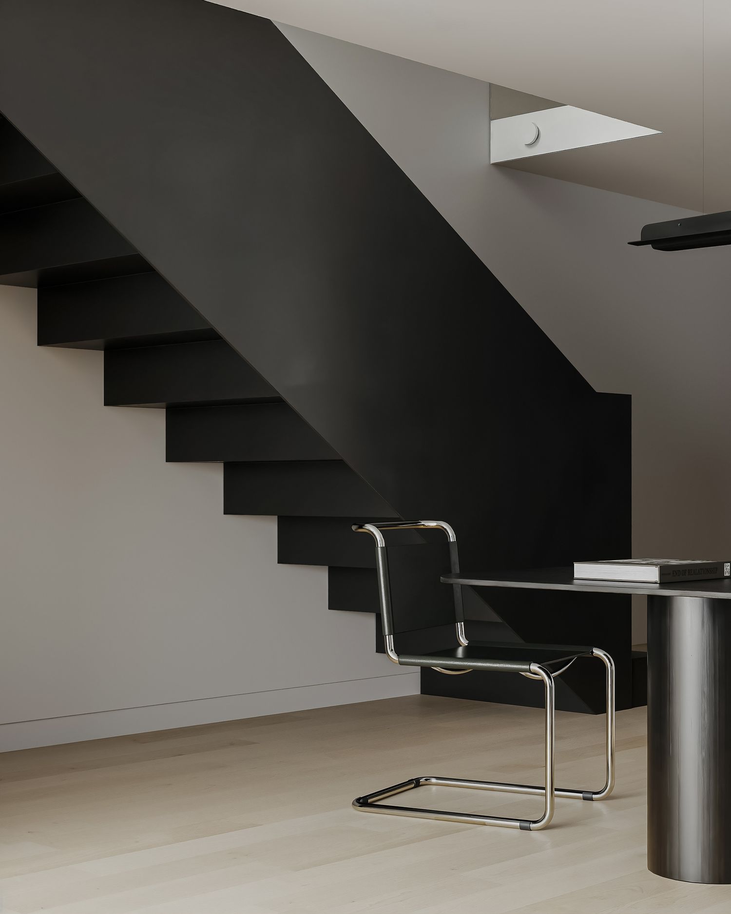 Modern minimalist interior featuring black floating staircase with chrome cantilever chairs and cylindrical table.